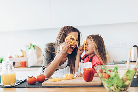 A mother and her young daughter laugh together as look through bell pepper rings while cutting vegetables for a salad.