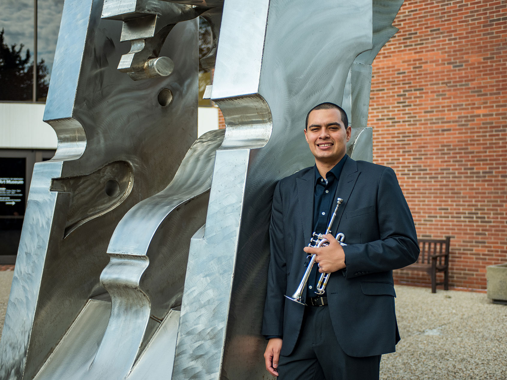 Adiel Nájera poses with his trumpet in front of an outdoor sculpture on the University of Kentucky campus.