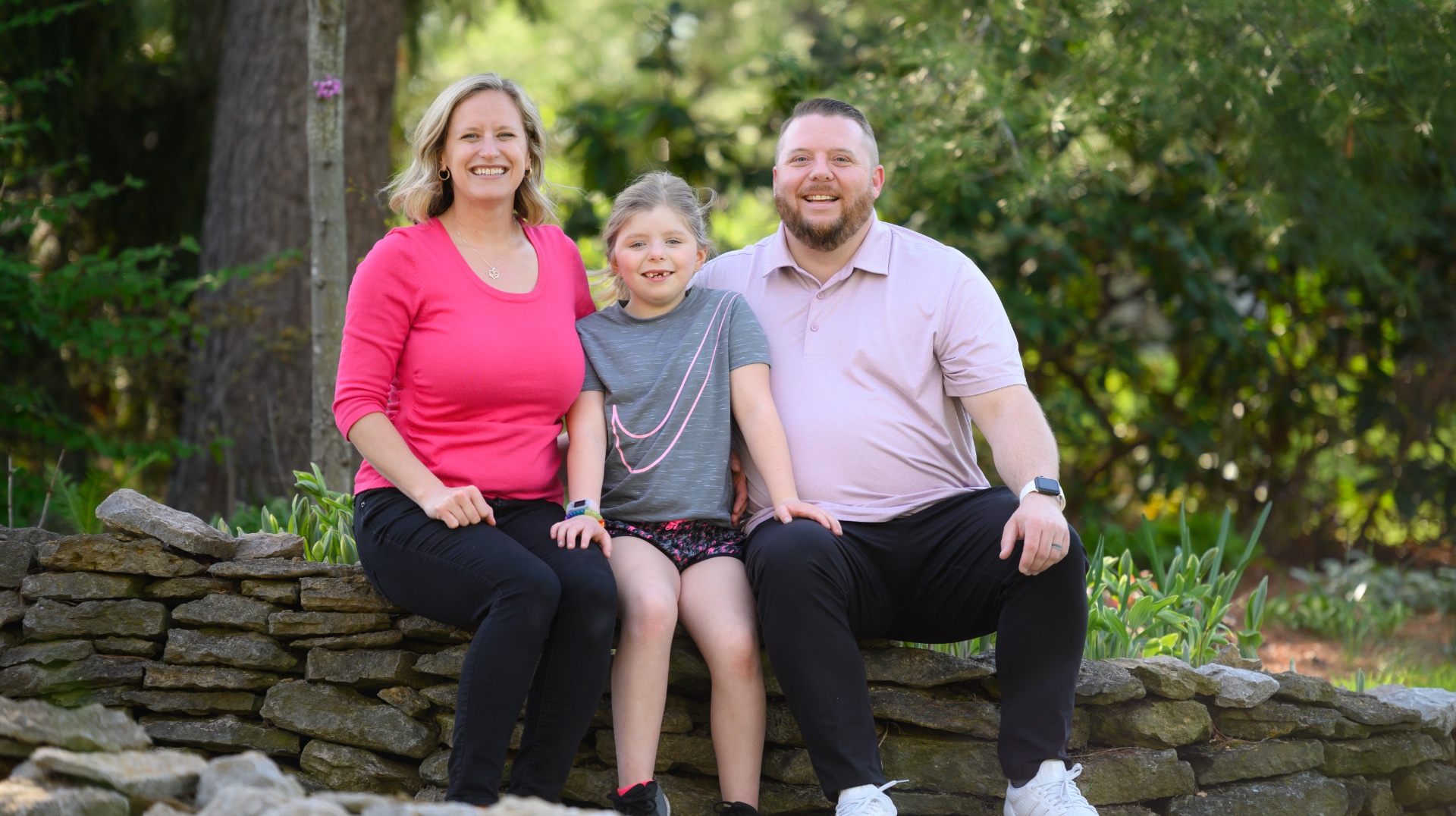 The Ferguson family sits outside and smile together. Sarah Beth is seated between her father, Scott and her mother, Erin, a middle-aged white woman with blonde hair, who is wearing a pink shirt.