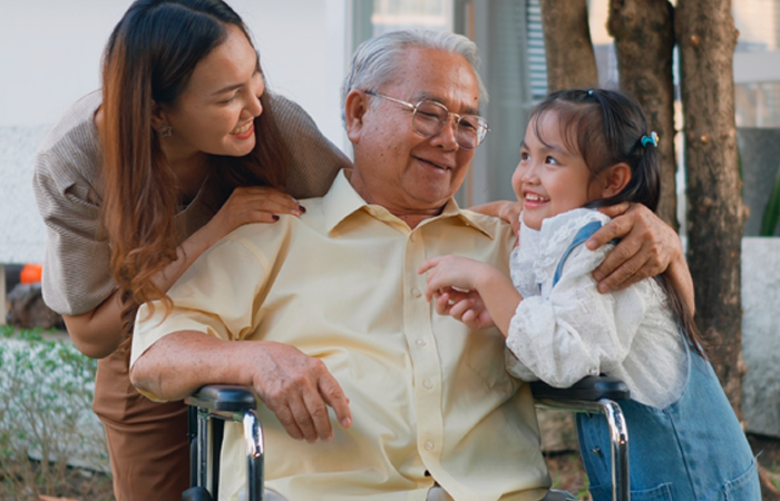 A daughter and granddaughter stand on either side of an older man who is wearing a yellow shirt and is seated in a wheelchair.