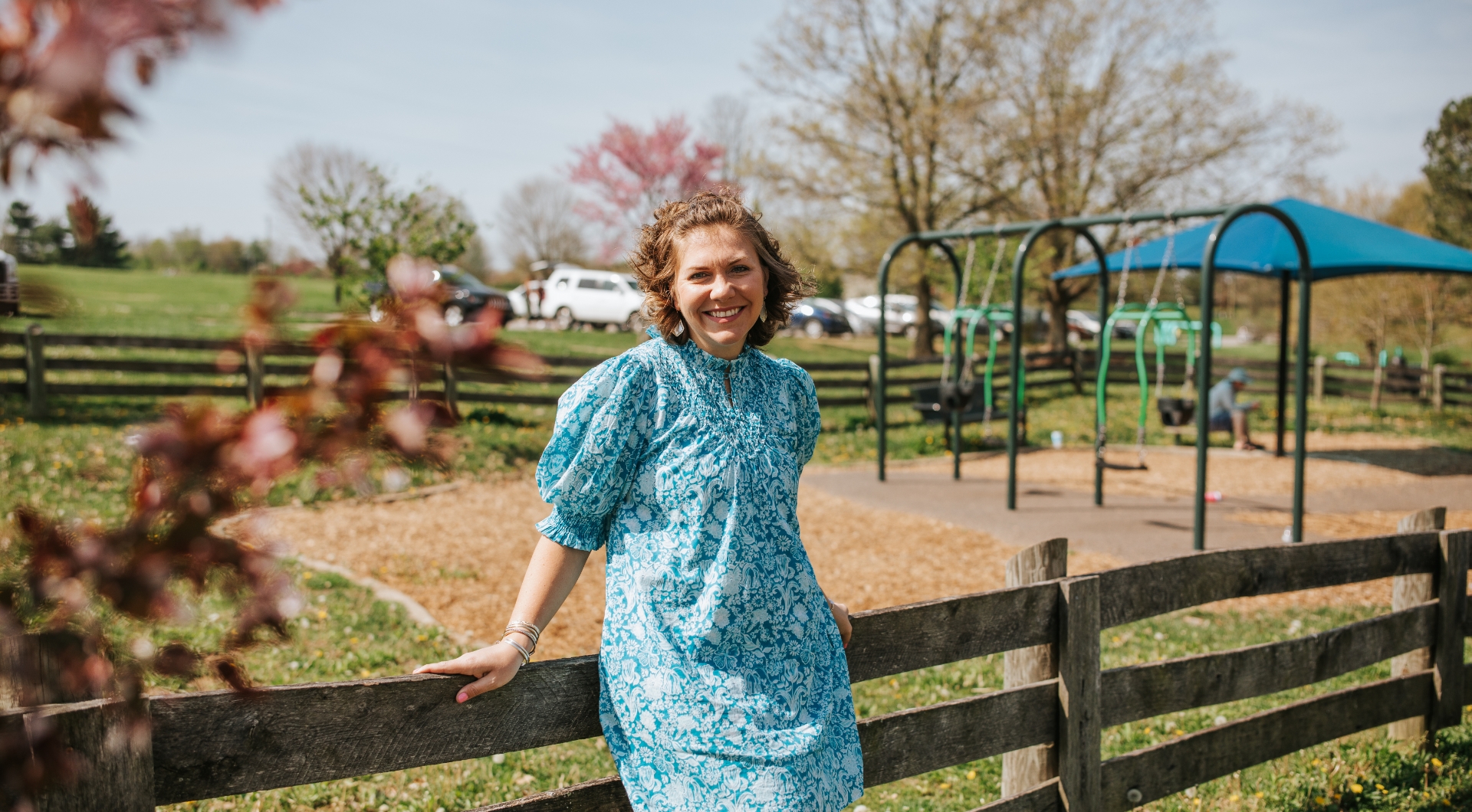 A picture of Anne Sydney, a white woman with short curly brown hair, smiling for the camera as she leans on a wooden fence at a children’s playground. She is wearing a white and turquoise dress that has puff sleeves and an intricate floral pattern.