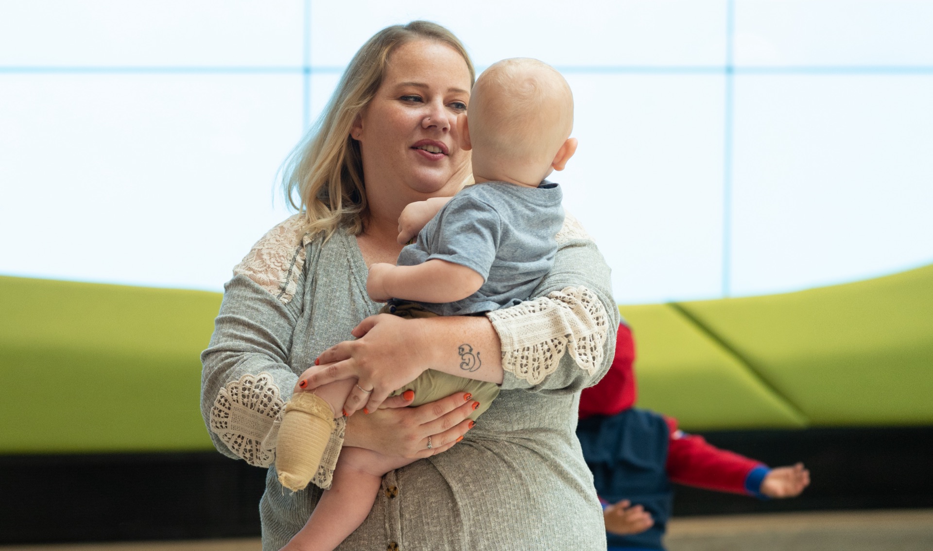 Emily Yost, a blonde woman wearing a grey shirt with white lace detailing, holds her son Arlo as he looks behind her.
