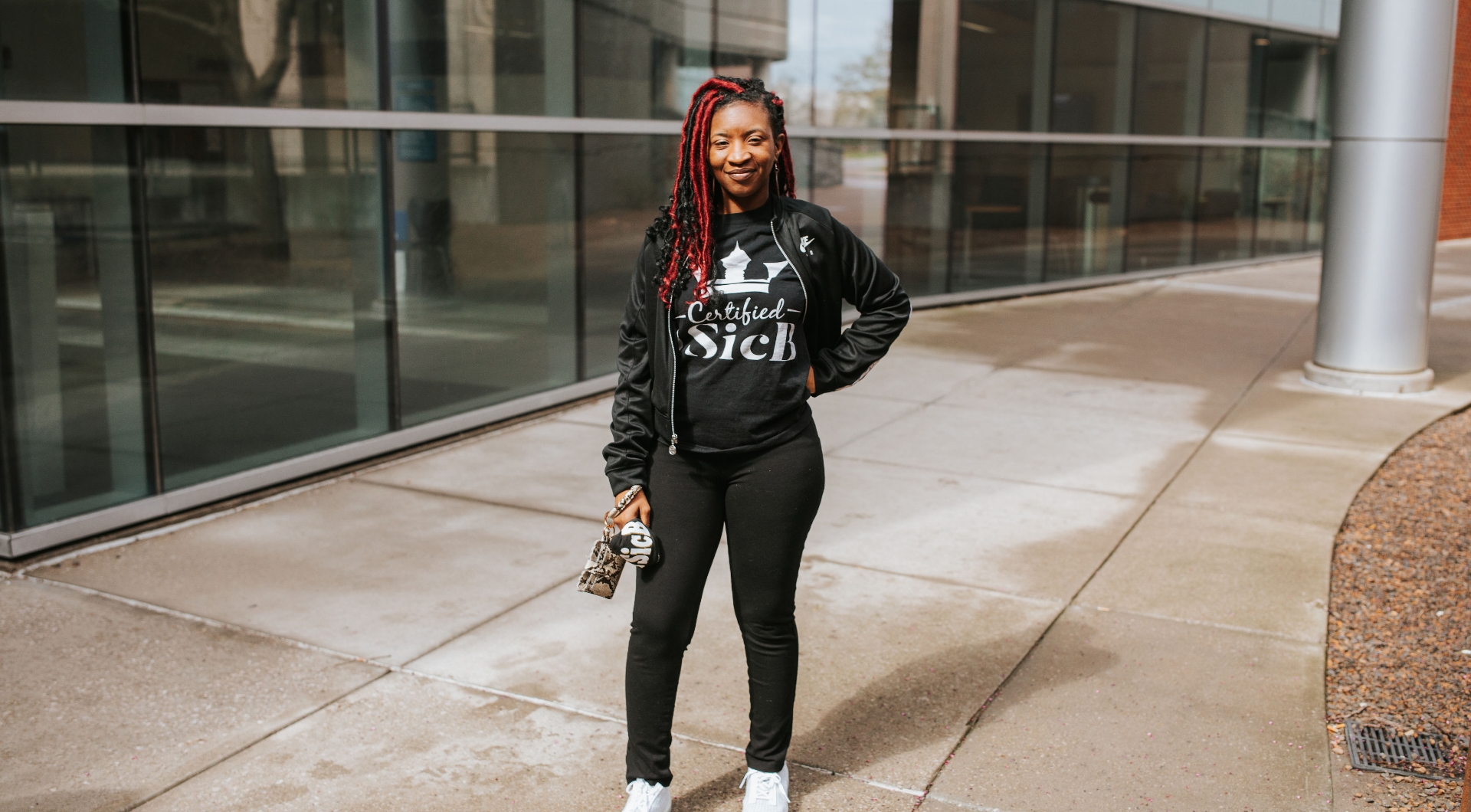 Brandi Jones, a Black woman with long red and black dreadlocks, poses for the camera with her hand on her hip. She is wearing a black Nike jacket, a black t-shirt that reads “Certified 1 SicB”, black pants, and circular earrings that read “SicB” in the middle.