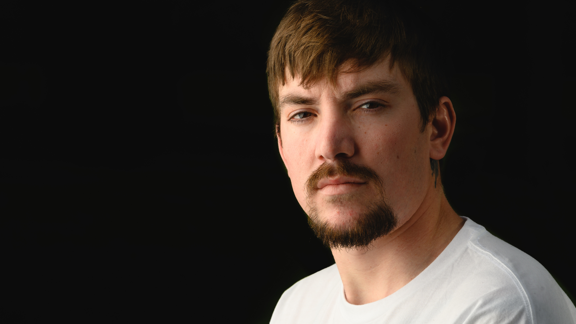 Cameron Waters, a white adult male with brown hair, wearing a white shirt stares straight into the camera with a black background.