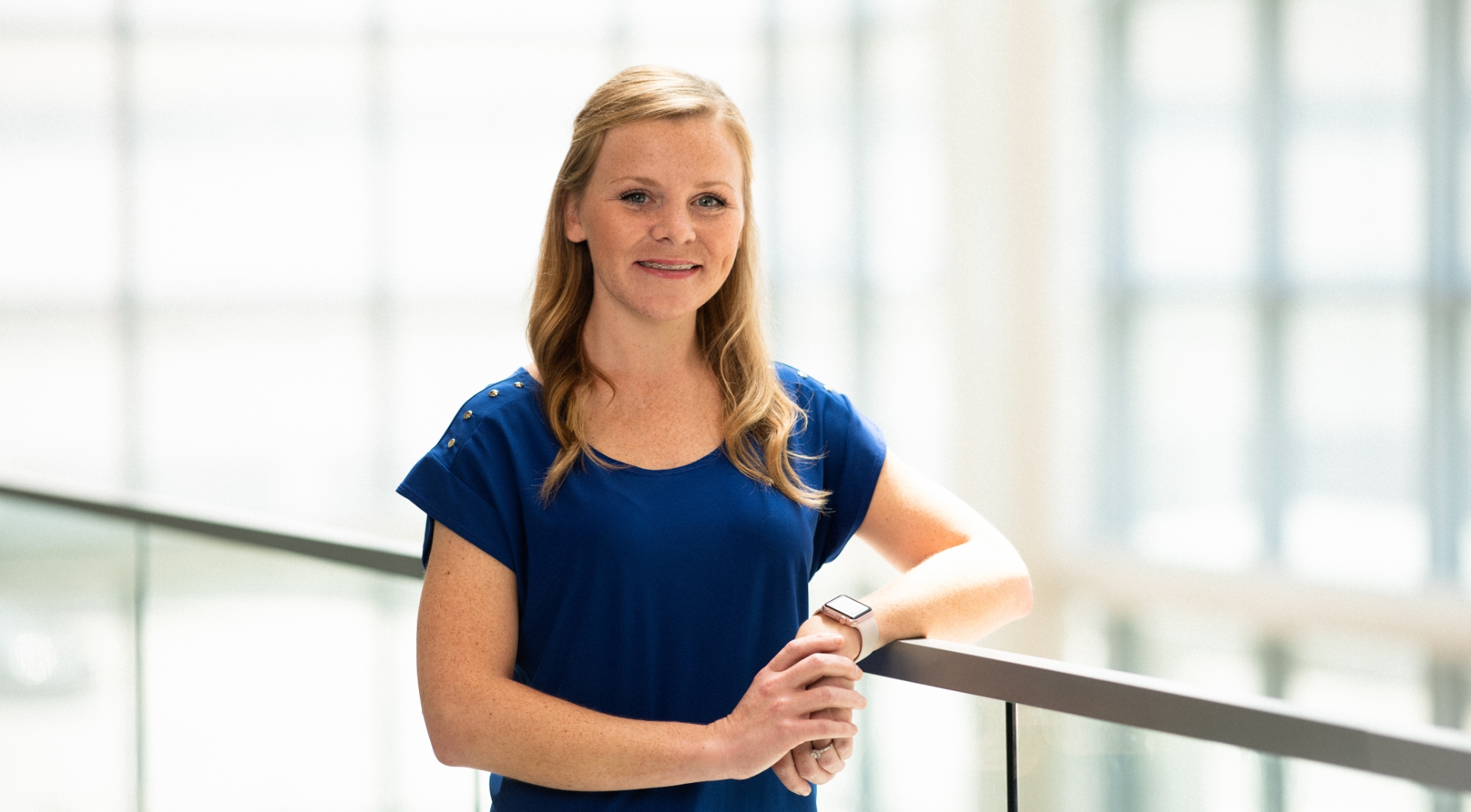 Christa Childers, a young white woman with long strawberry-blonde hair, leans on a rail while smiling for the camera. She is wearing a navy blue shirt that has button detailing on the shoulders, and an Apple watch on her wrist.