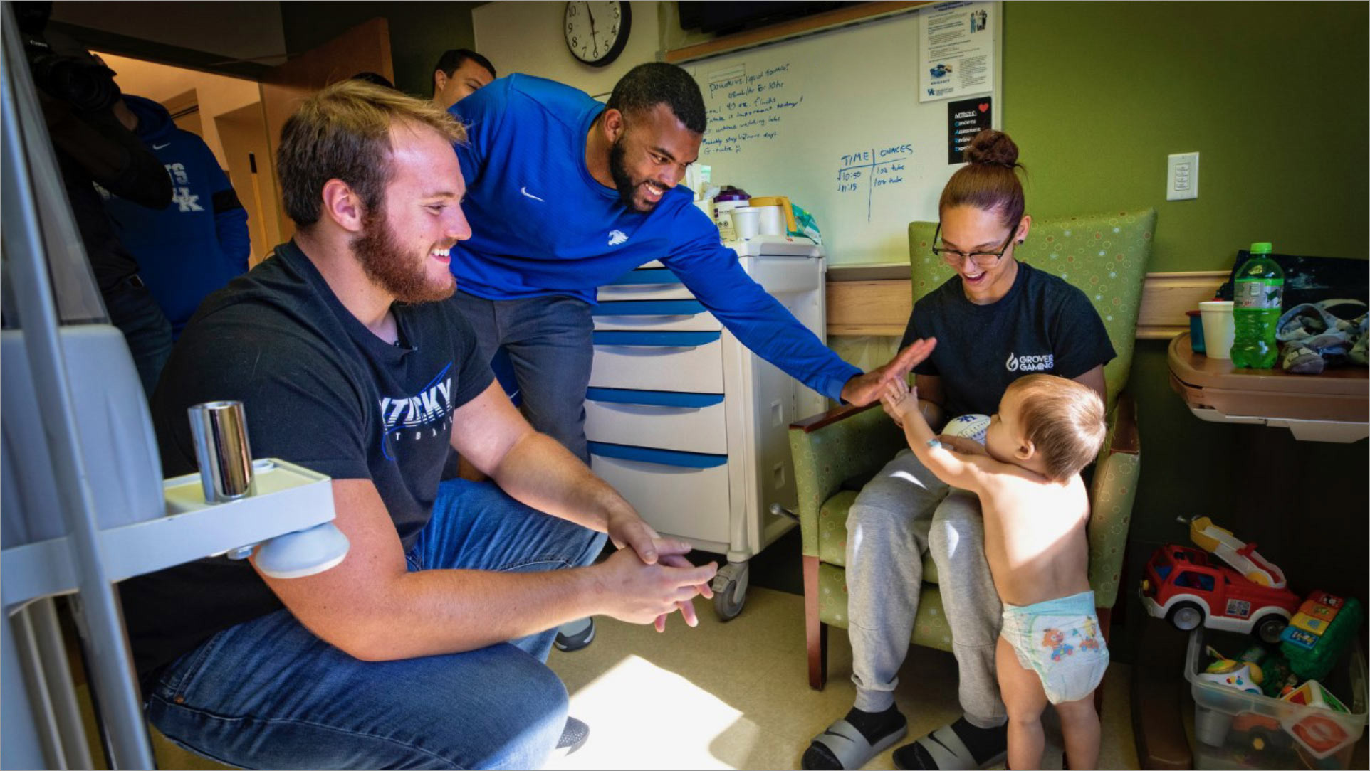 Courtney Love bends down to give a toddler a high five. There are several other football players in the room, along with the toddler’s mother.