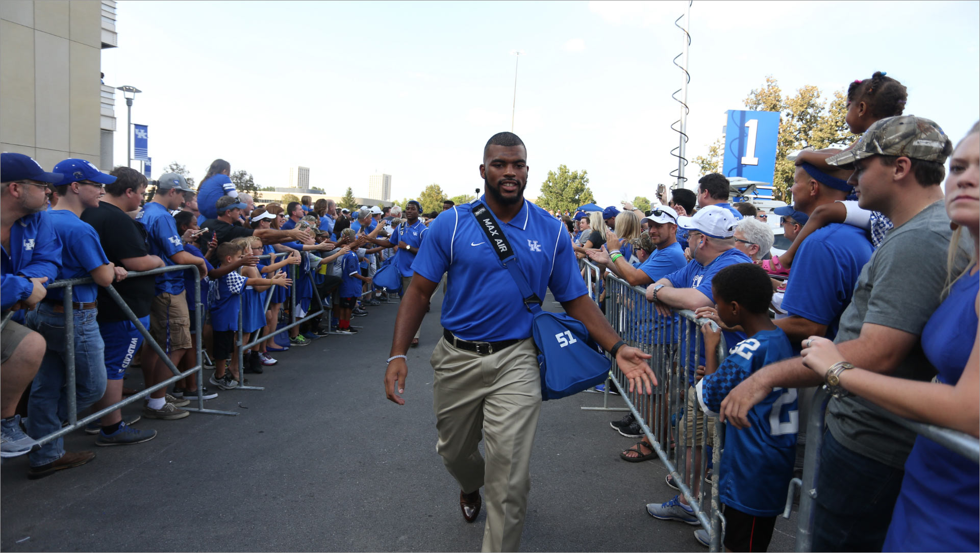 Courtney, wearing a blue polo shirt, walks down the center of two barriers with UK fans behind them. He holds out his hand to give a little boy a high five.