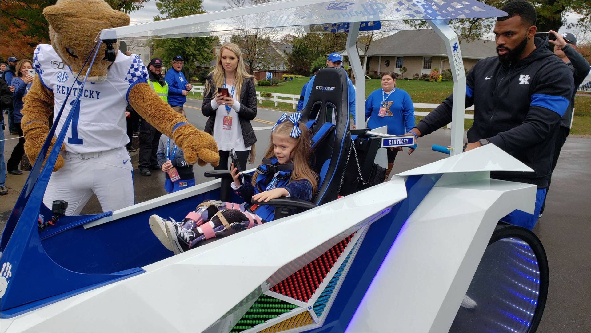 Courtney and the Wildcat mascot surround a little girl in a futuristic-looking blue and white cart. The girl has her hair in pigtails and has pink and purple leg braces.