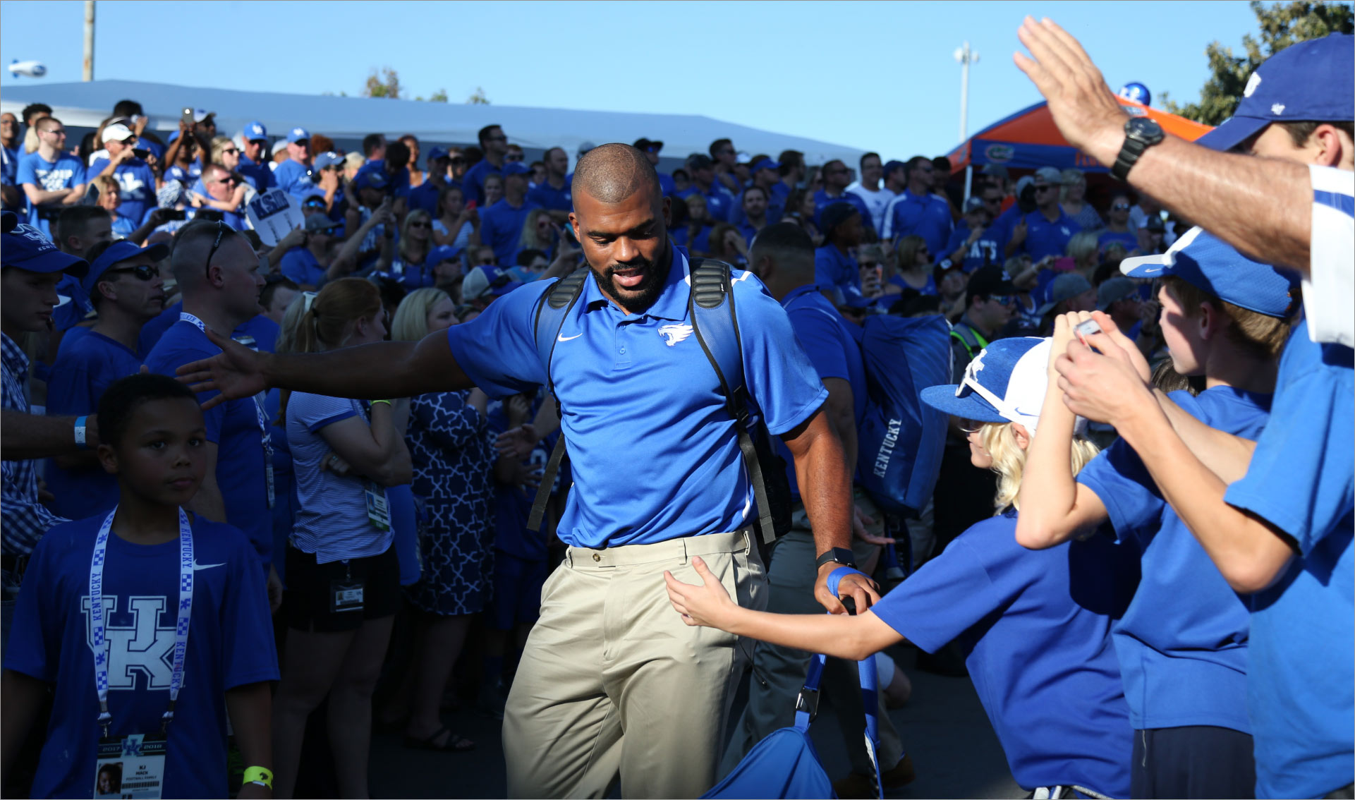 Courtney walks through a crowd of fans, all wearing blue, before a football game. He holds his arm out to give a kid a high five.
