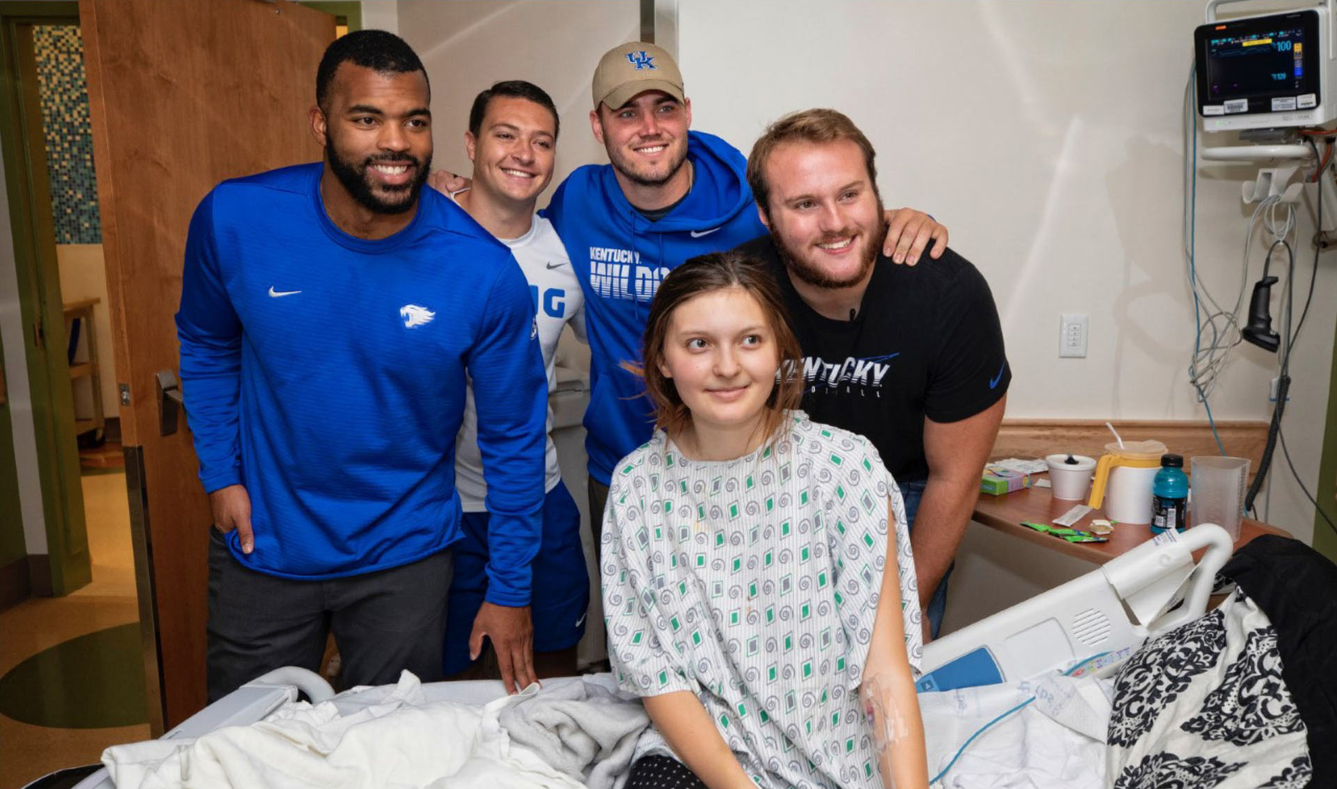 Courtney Love, wearing a blue shirt, and three current football players wearing Kentucky gear, pose with a Kentucky Children’s Hospital patient. The patient, a young girl, is sitting in her bed and wearing a hospital gown.