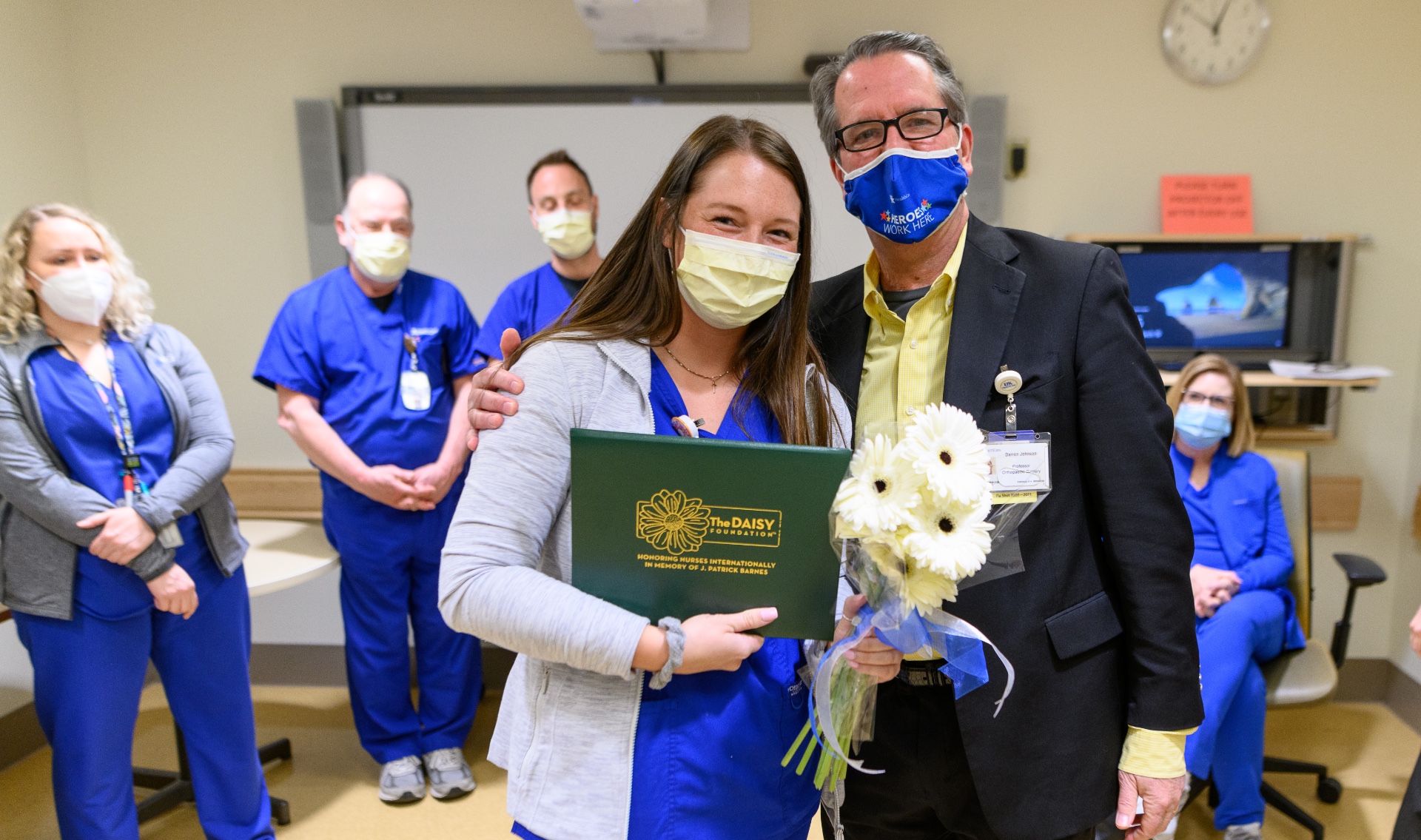 Holding a bouquet of flowers, Alexis Zody, an adult white woman with brown hair in blue scrubs, stands beside Dr. Johnson for a photograph. They are surrounded by colleagues, and everyone pictured is wearing a facemask.