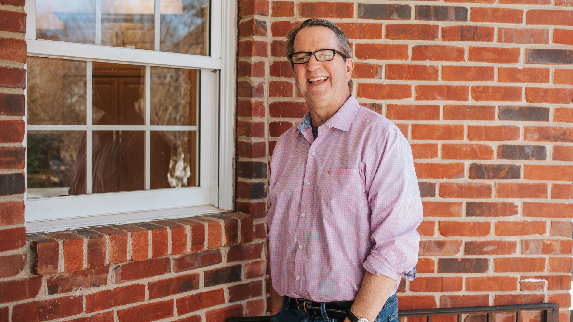 Dr. Darren Johnson, an older white man with black and grey hair, stands outside in front of a brick wall. He is wearing a light pink checkered shirt, blue jeans and black spectacles.