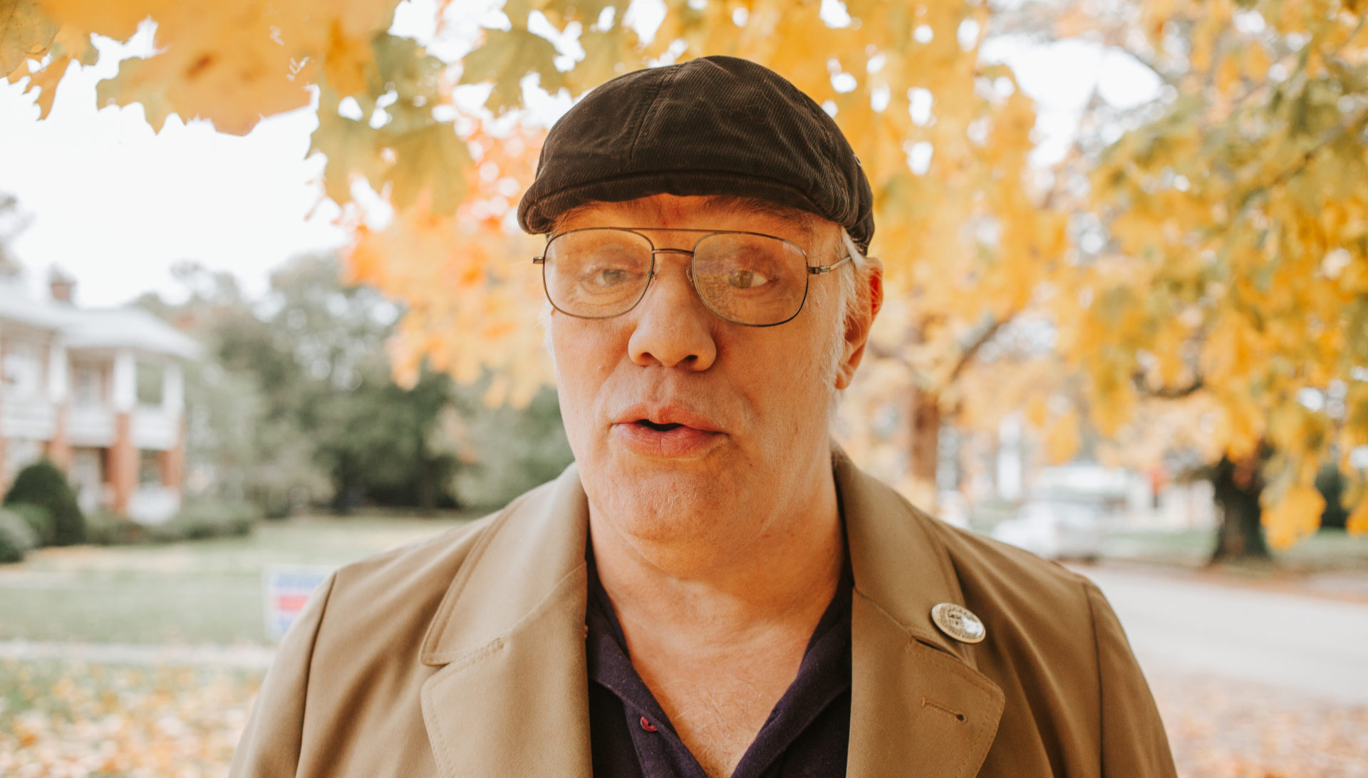 Donald Young, an elderly white man, stands in an outdoor setting under a tree. He is wearing a black ivy hat with black-framed spectacles and a brown coat.