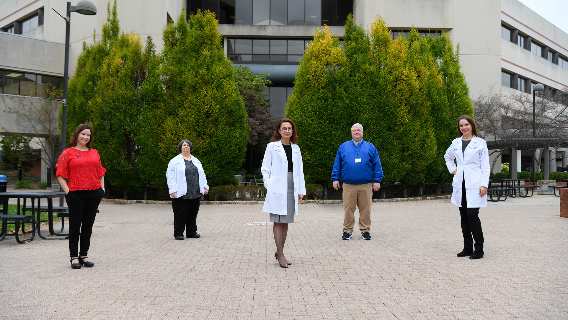 Dr. Meriem Bensalem-Owen, a middle-aged white woman with red hair, stands in an outside setting area with two of her colleagues: a middle-aged white man and an elderly white woman. Dr. Bensalem-Owen is wearing a white doctor's coat, a grey skirt and black framed glasses with a black blouse.