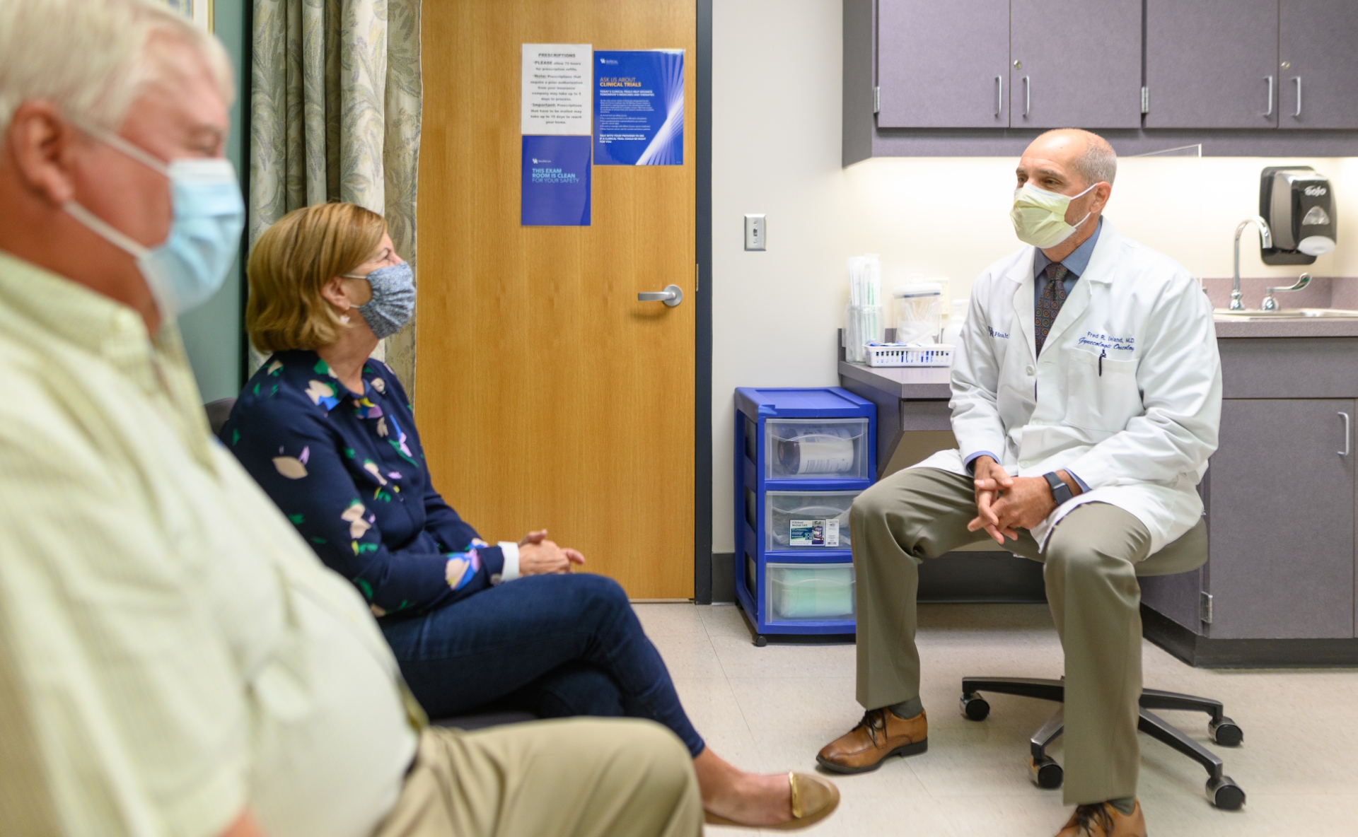 E and her husband Cary, an older white man with white hair, sit in a hospital room with Dr. Fred Ueland, a middle-aged white man with short-cropped grey hair. Dr. Ueland is wearing a white lab coat. All three are wearing masks.