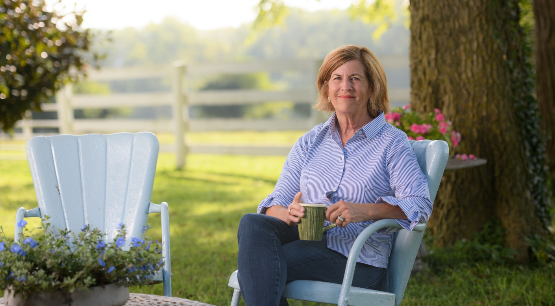 E Barr, an older white woman with shoulder-length light brown hair, sits in a lawn chair holding a cup of coffee.