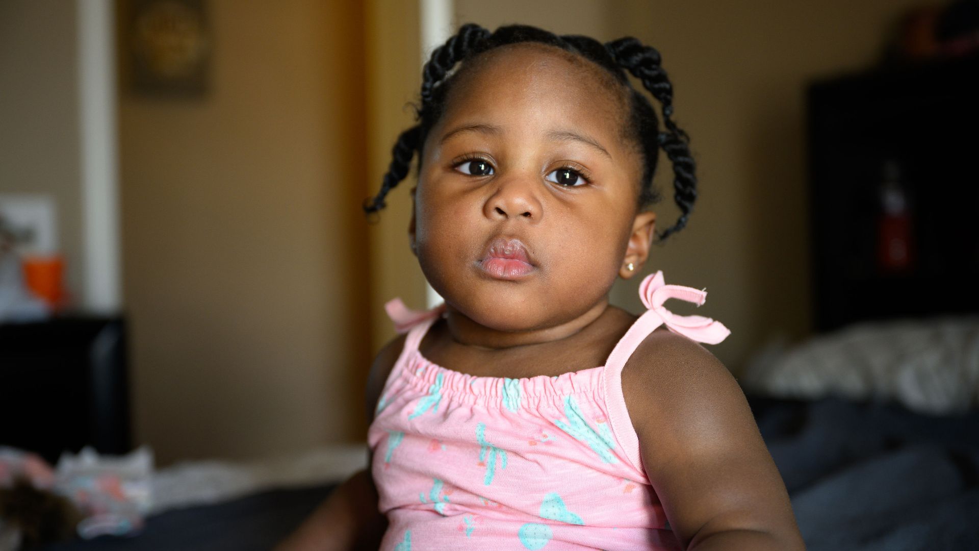 A photo of Elizabeth (Betty) Kargbo sitting on a bed as she looks into the camera. She is a young Black toddler with dark hair in twists. She is wearing a pink and blue top that is tied at the shoulders.