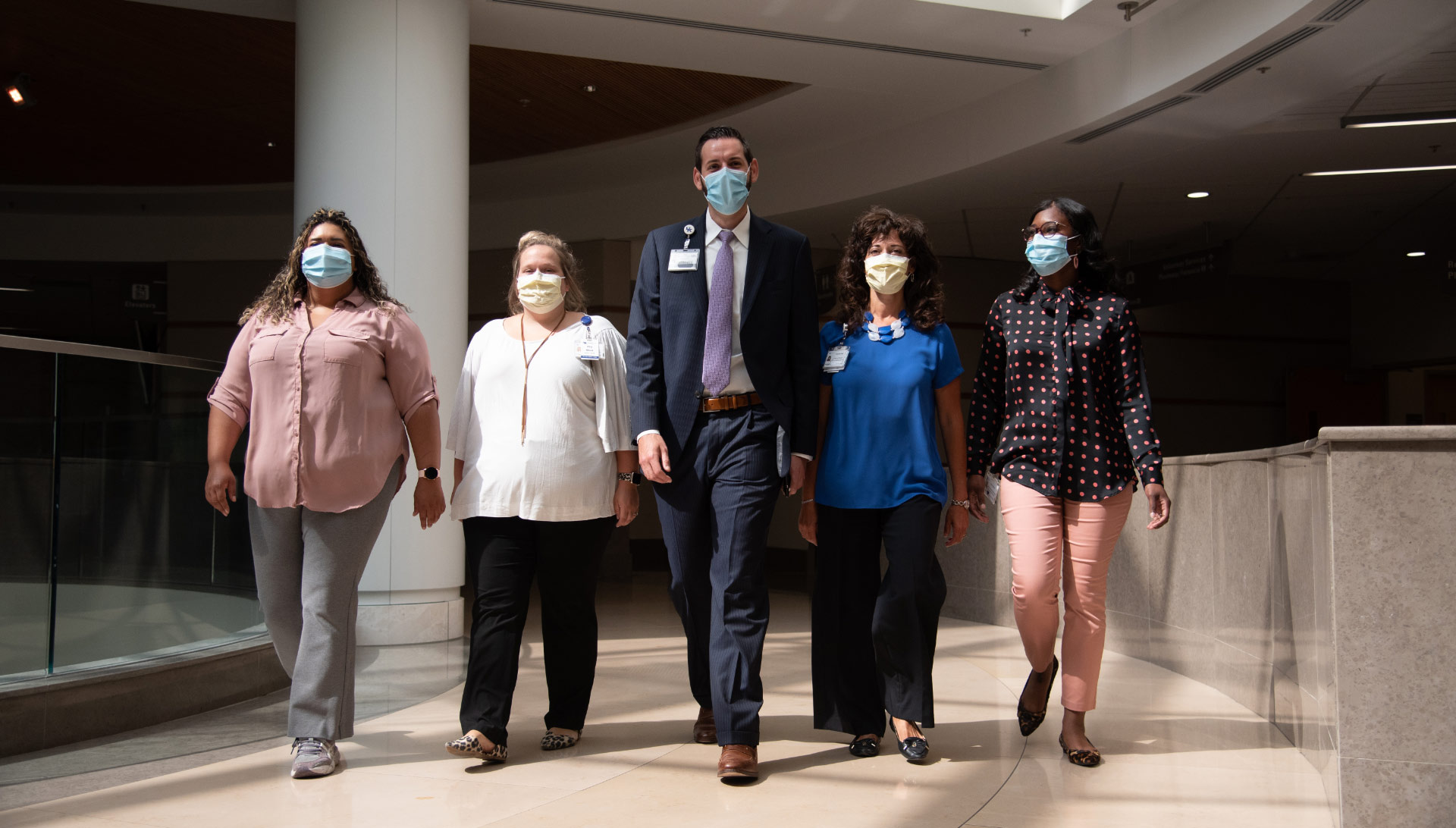 A group of people walking side by side down an open hallway, all wearing face masks.