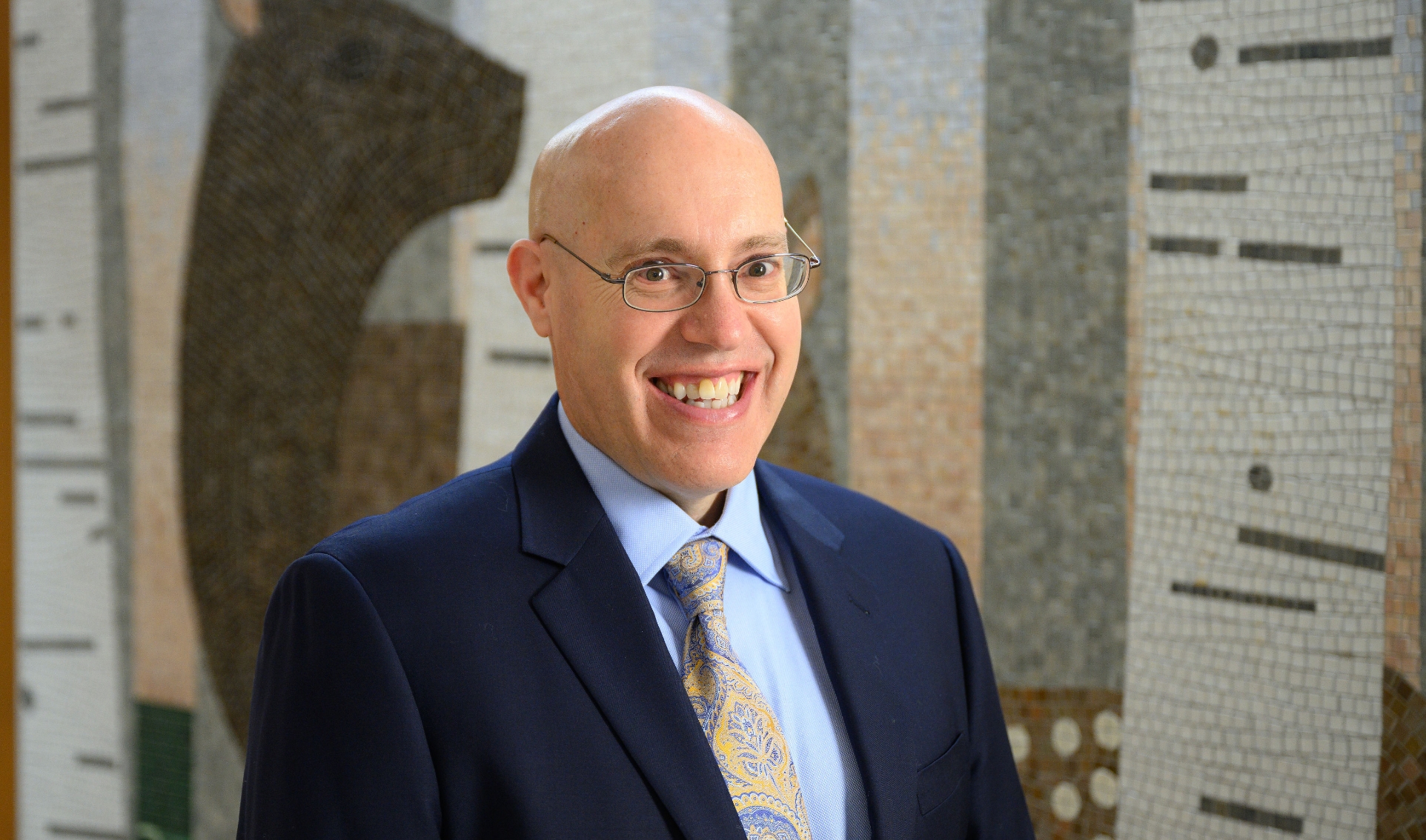 A photo of Dr. Hubie Ballard enthusiastically smiling while standing in front of a mosaic. He is a white bald man who is wearing glasses, a navy suit jacket, a light blue shirt, and a paisley tie.