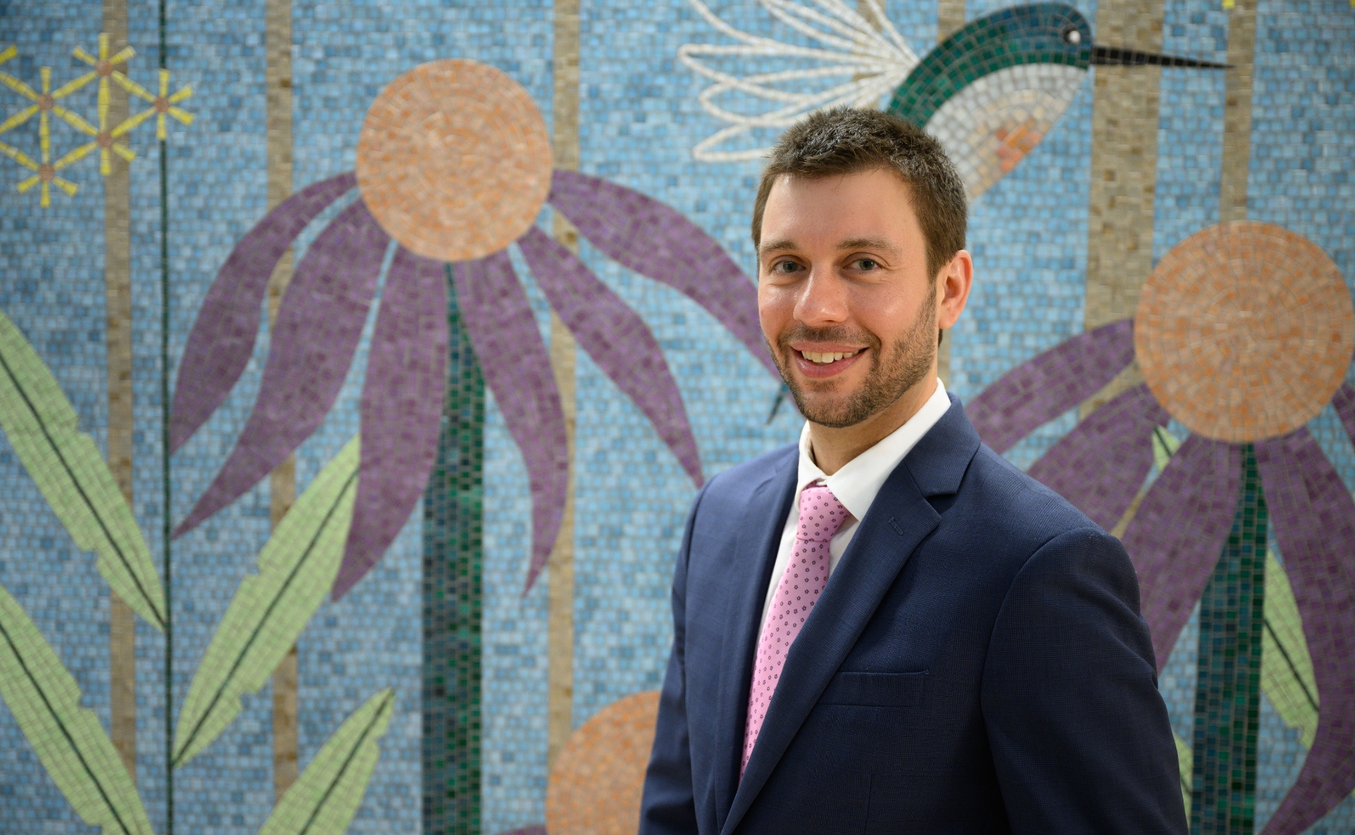 A photo of Dr. Kenneth Iverson smiling as he stands in front of a mosaic of a hummingbird and flowers. Dr. Iverson is a young white man with short brown hair. He is wearing a navy suit jacket with a white button-up shirt and a pink tie.