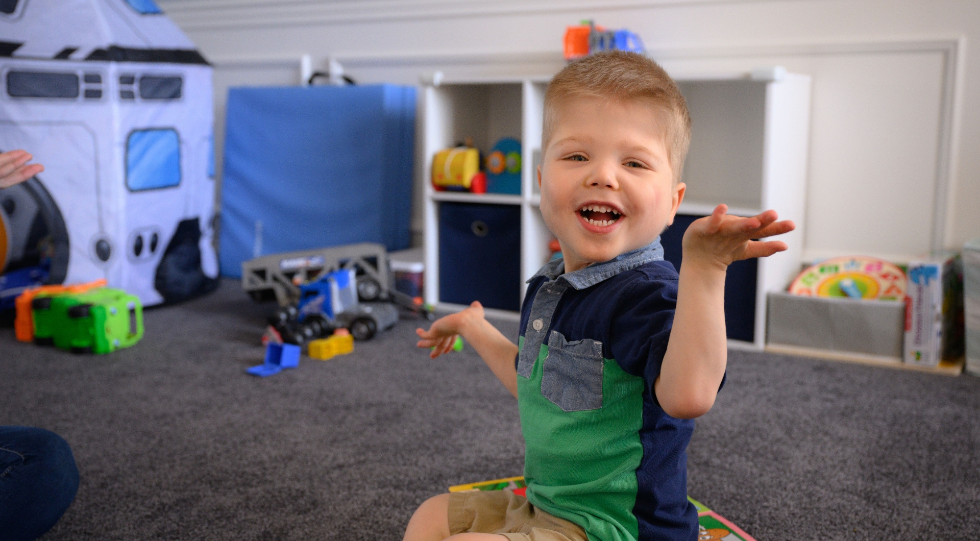 A candid photo of Everett Polston, a white male toddler with short blonde hair, gesturing with his hands as he plays with his toys. He is wearing a half green half blue short-sleeve polo, and a pair of khaki shorts.