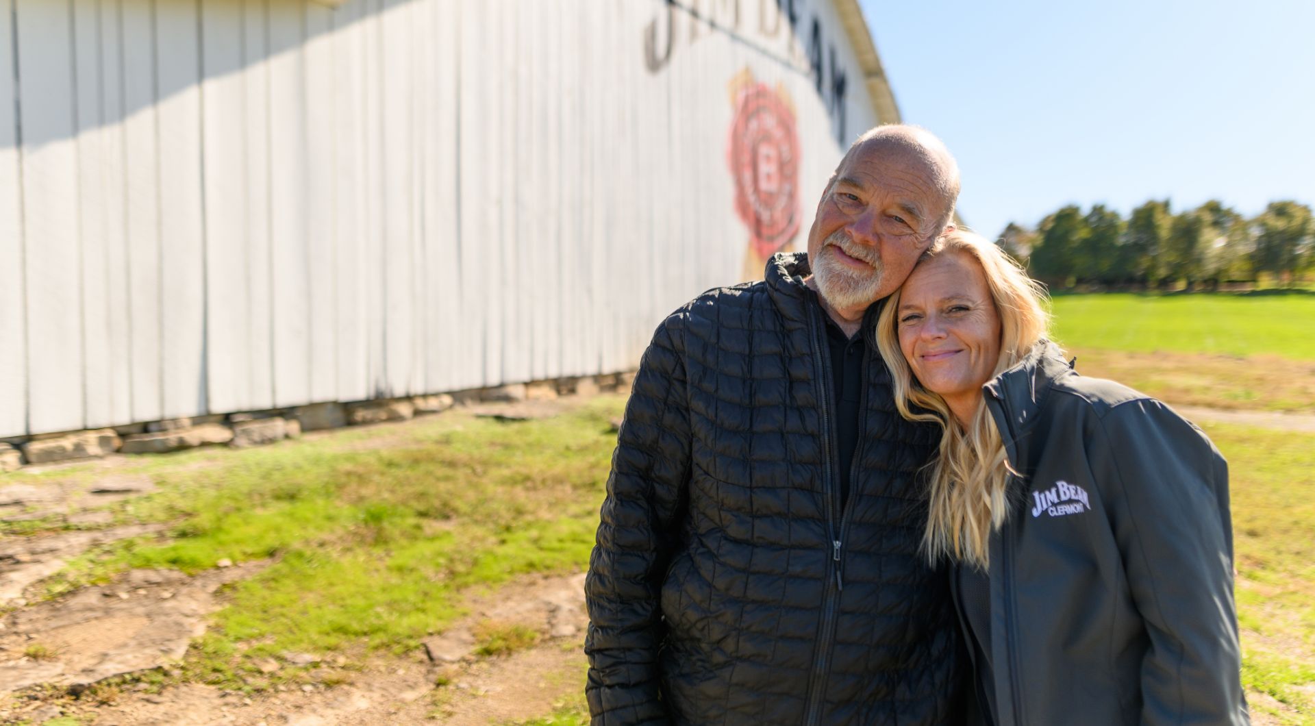 Fred Noe, an older white man with a bald head and a gray goatee, poses outside at a James B. Beam Distillery with DeeAnn Hogan. She is an older white woman with long blonde hair, wearing a gray Jim Bean zip-up jacket. He is wearing a black puffer jacket over a navy t-shirt.