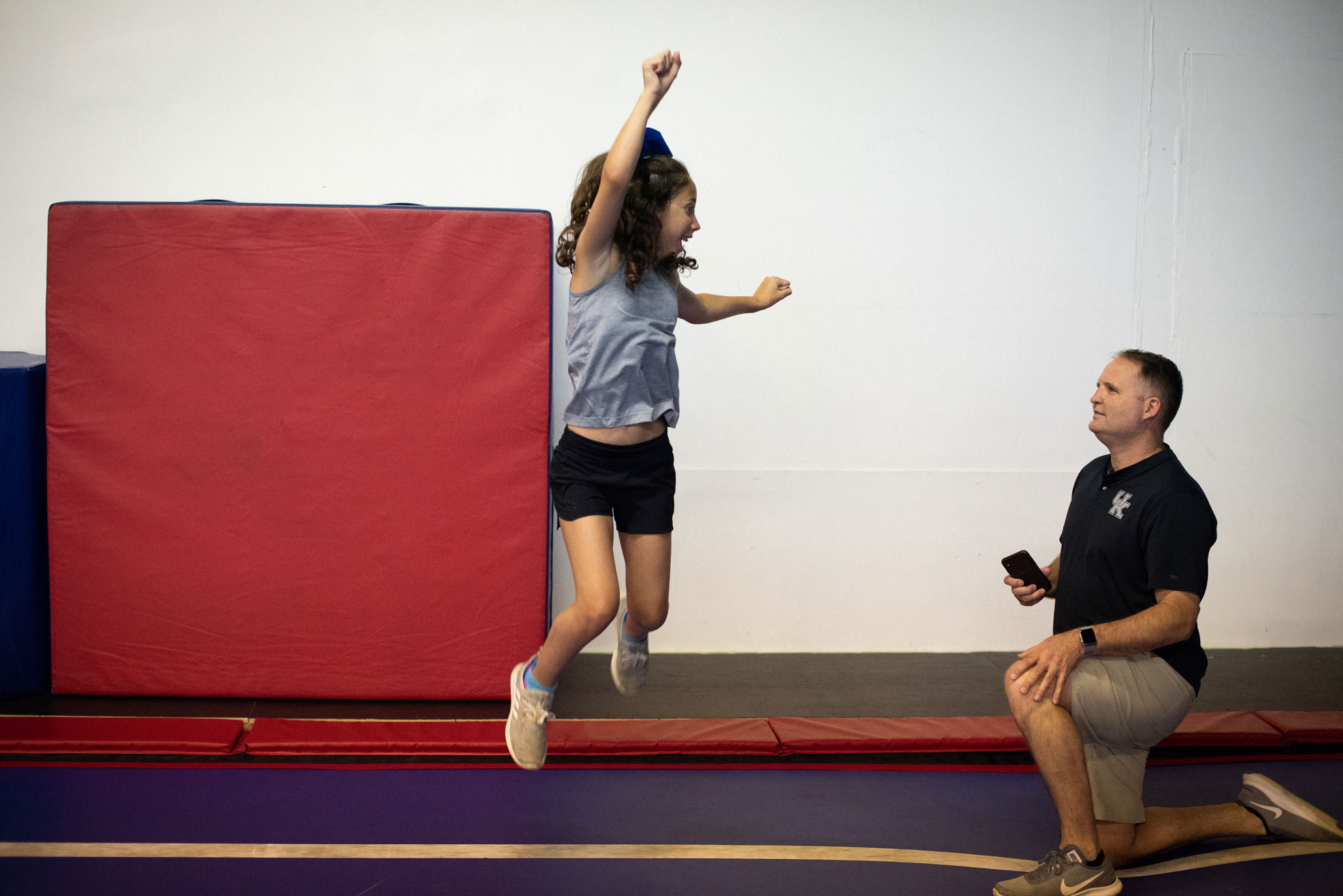 Gracie jumps enthusiastically on a gymnastics mat while her father, a middle aged man in a black polo shirt, watches her.