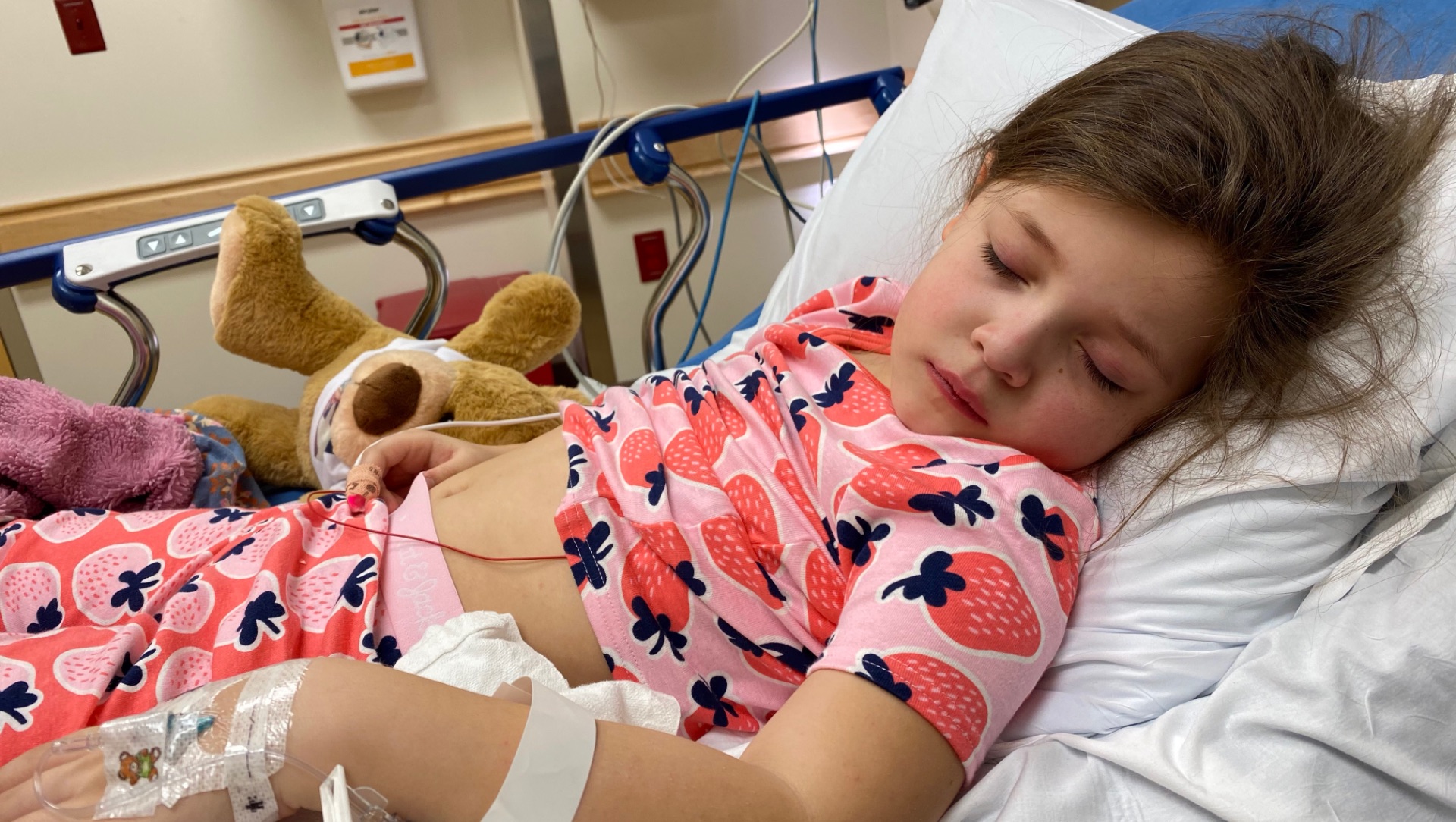 Henley lays in a hospital bed with different wires attached to her. She is wearing pink pajamas with strawberry designs on them.