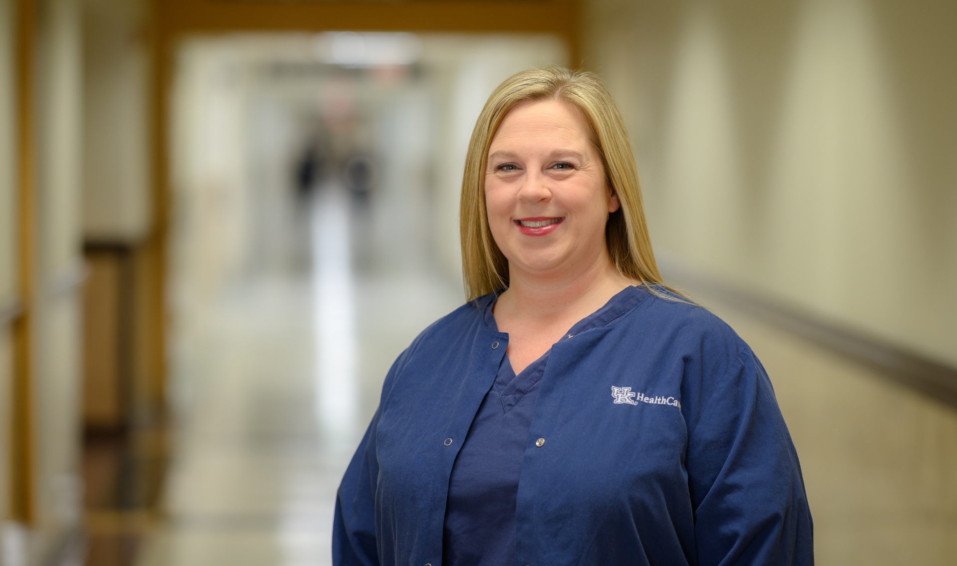 Allison Pinkston, a middle aged blonde white woman, stands smiling in the hospital hallway. She is wearing blue scrubs, as part of the required uniform.