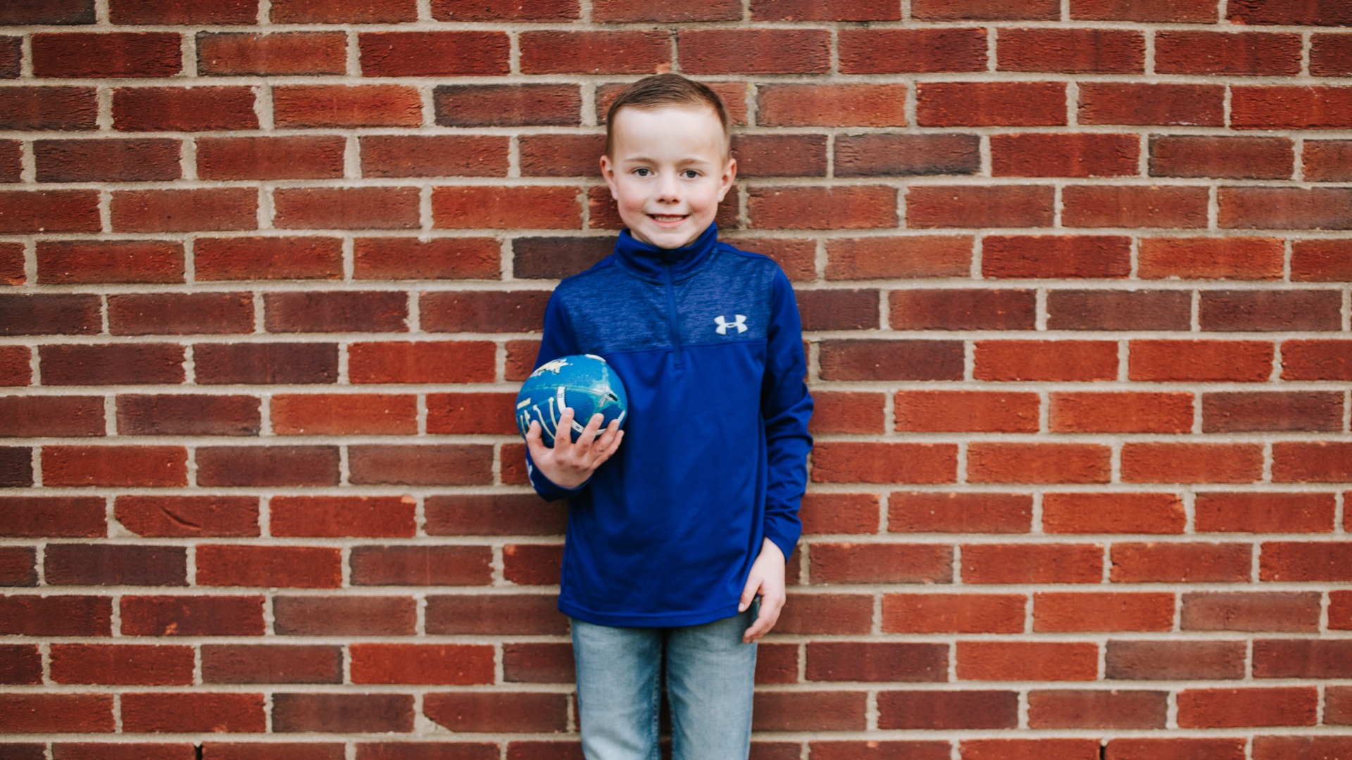 Henry Longeway, a young white boy with brown hair, stands in front of a brick wall, holding a blue football. He smiles, wearing a blue sweatshirt with blue jeans.