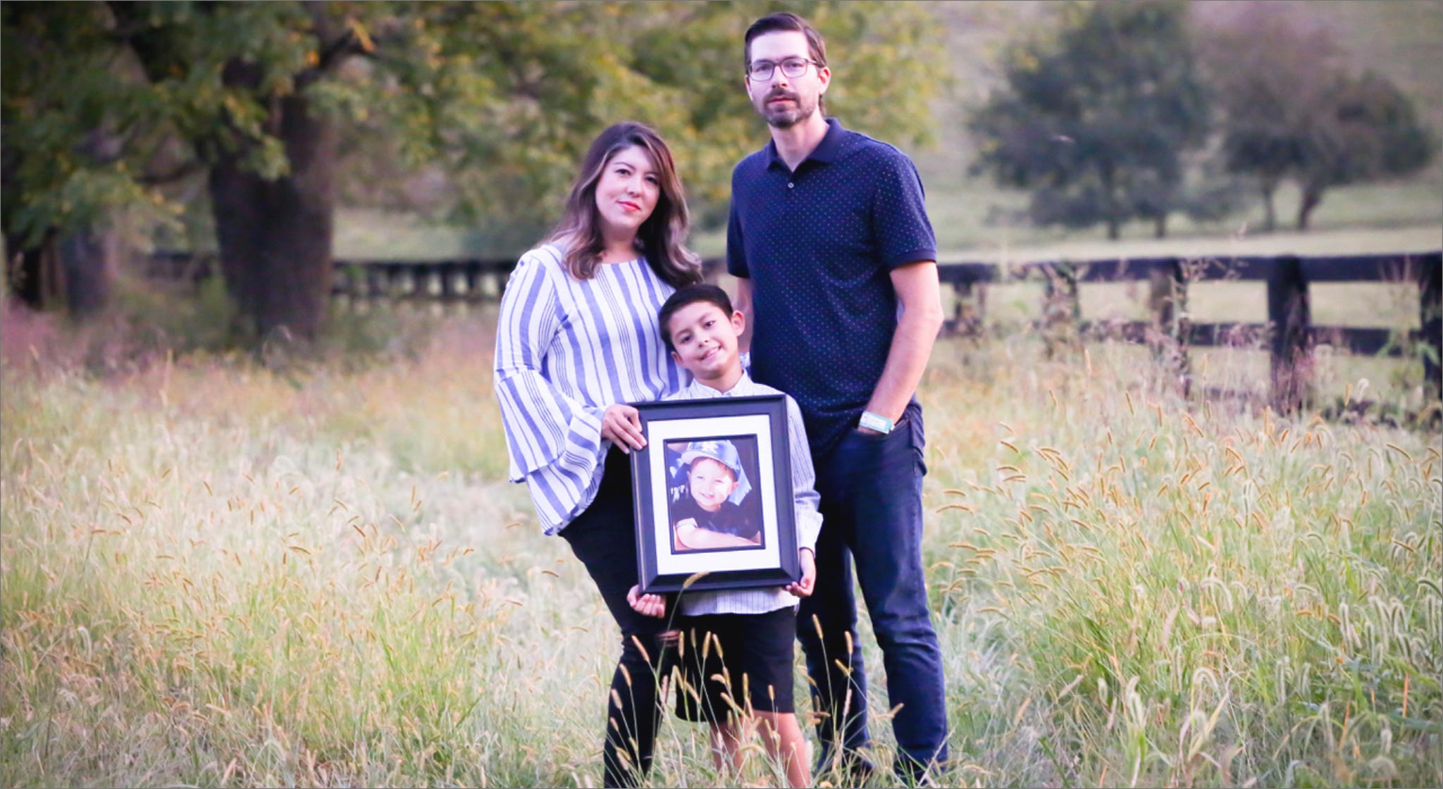 Liz, Benjamin, and Maximo stand together in a field for a family portrait. Maximo holds a large, framed portrait of Marco.