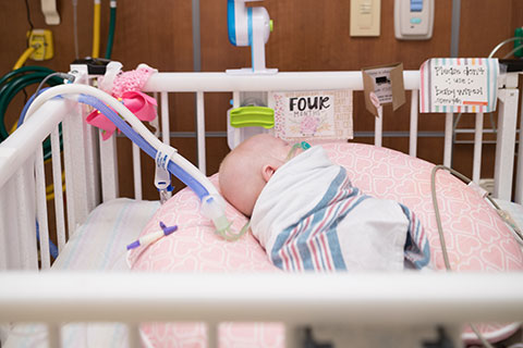 A newborn swaddled in a white blanket with pink and blue stripes sleeps in an open crib in the NICU.