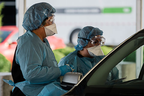 Two front-line workers wearing masks, goggles, and other protective gear work at a drive-thru COVID-19 testing site.