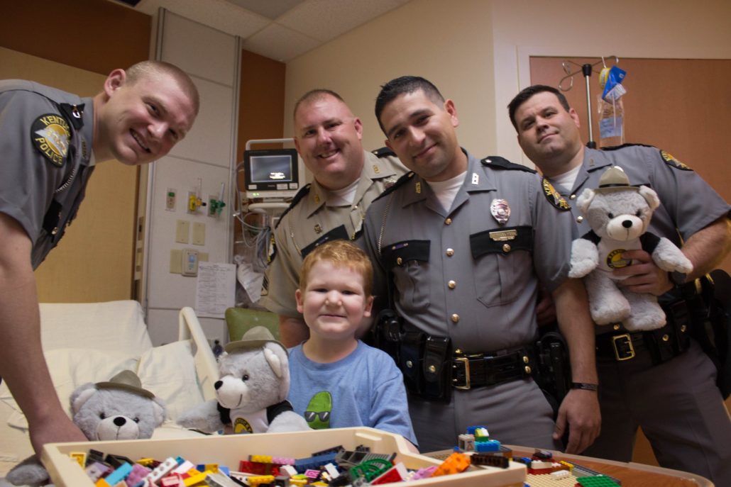 Four Kentucky State Troopers deliver a teddy bear to a young boy at Kentucky Children's Hospital.