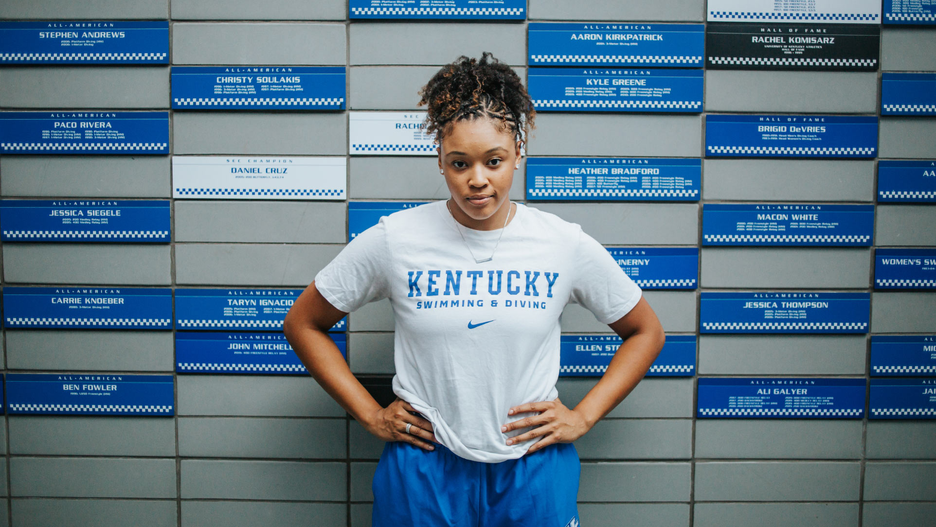 Jaida poses with her hands on her hips, wearing a white University of Kentucky t-shirt, in front of a wall with tiles of names of past University of Kentucky swimmers and divers.