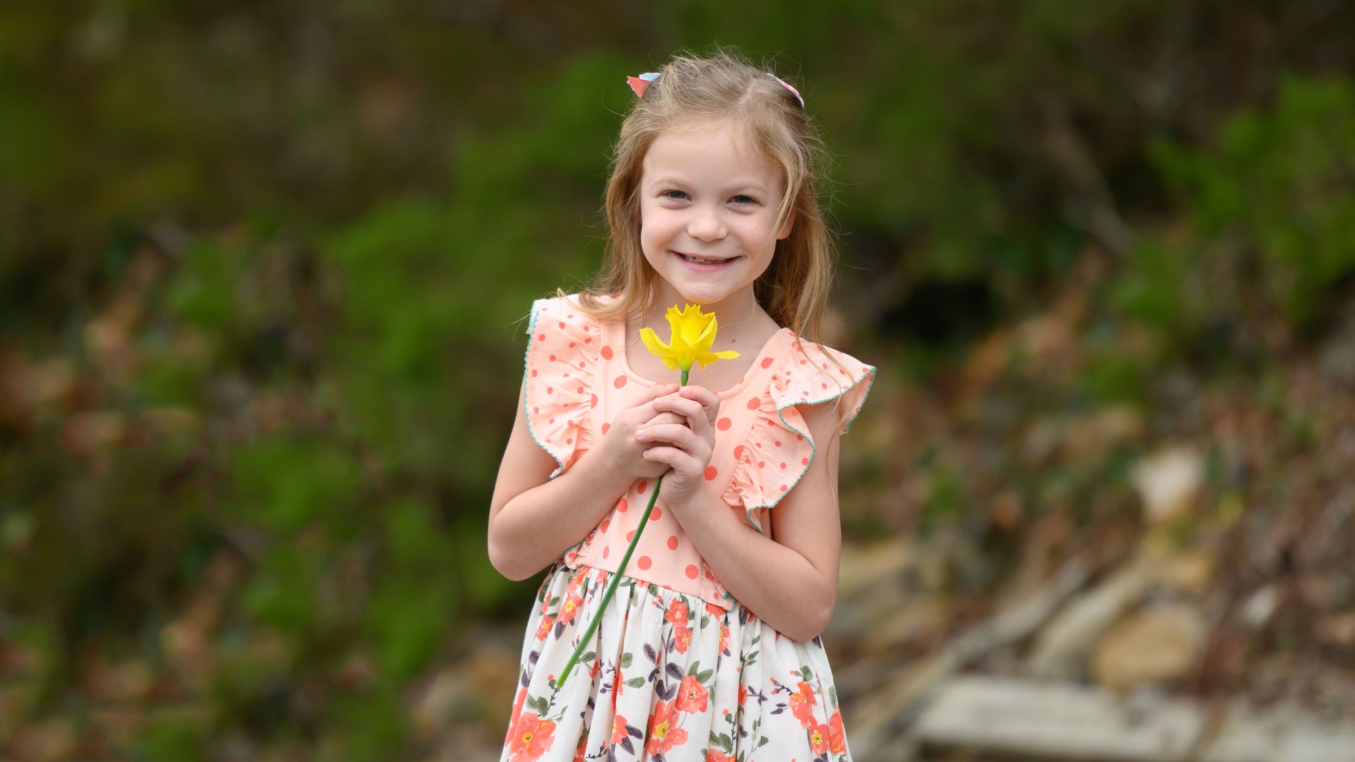 Julie Tate, a young white girl with blonde hair, stands smiling in an outside setting. She is wearing a white dress with orange polka dots and flowers while holding a yellow flower.