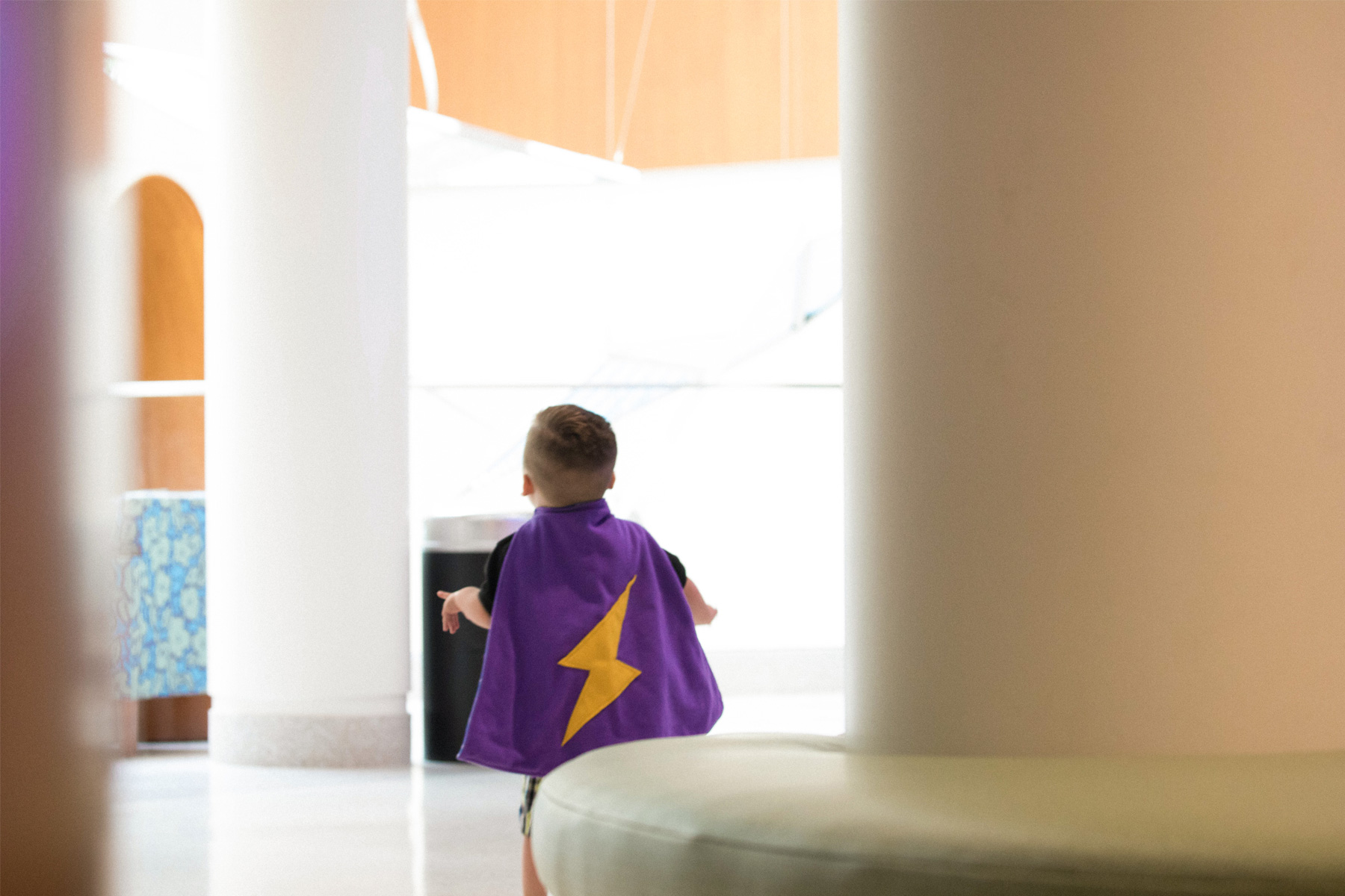 Kase is walking away wearing a purple cape decorated with a large yellow lightning bolt on his back.