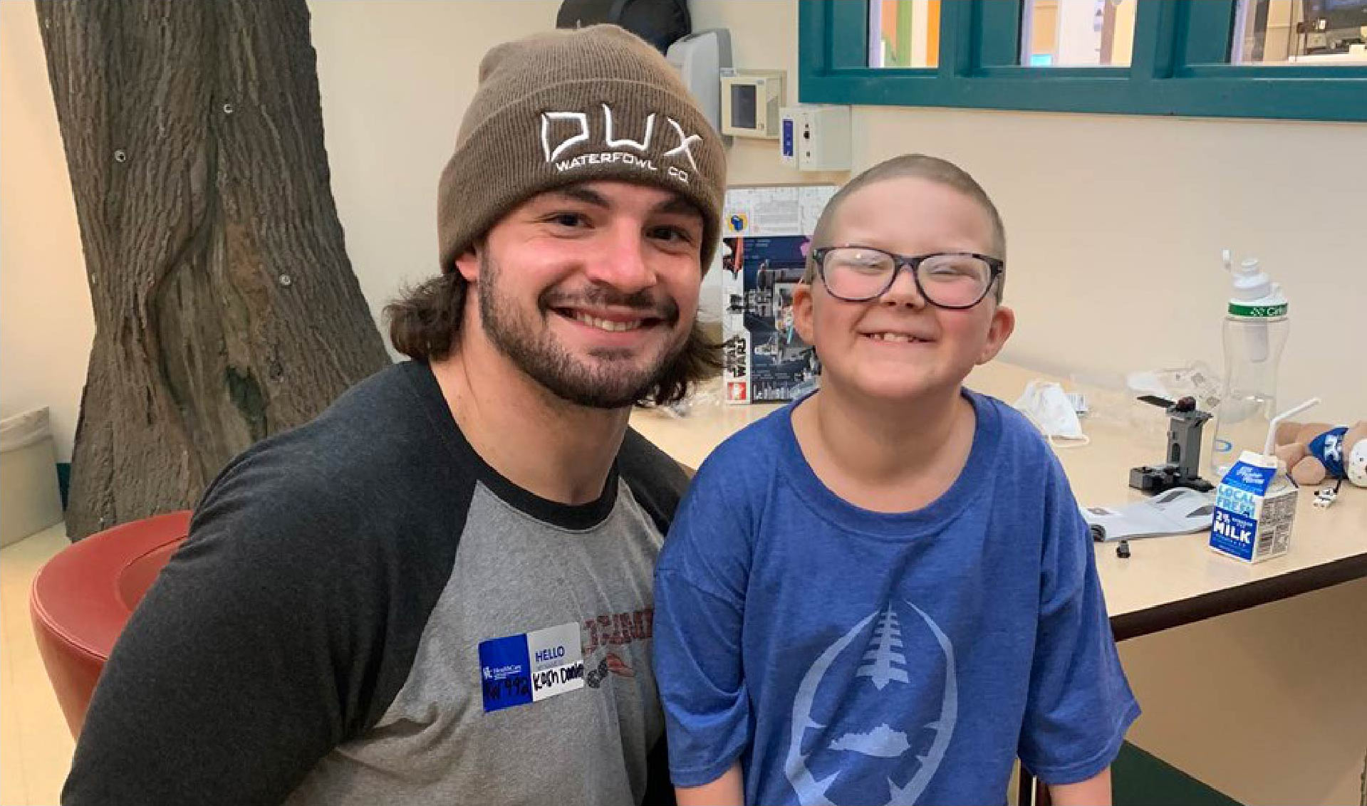 Kash Daniels, a football player, poses for a picture next to a little girl, Ellie. Ellie has buzzed hair and glasses. Kash is wearing a brown toboggan. Both are smiling.