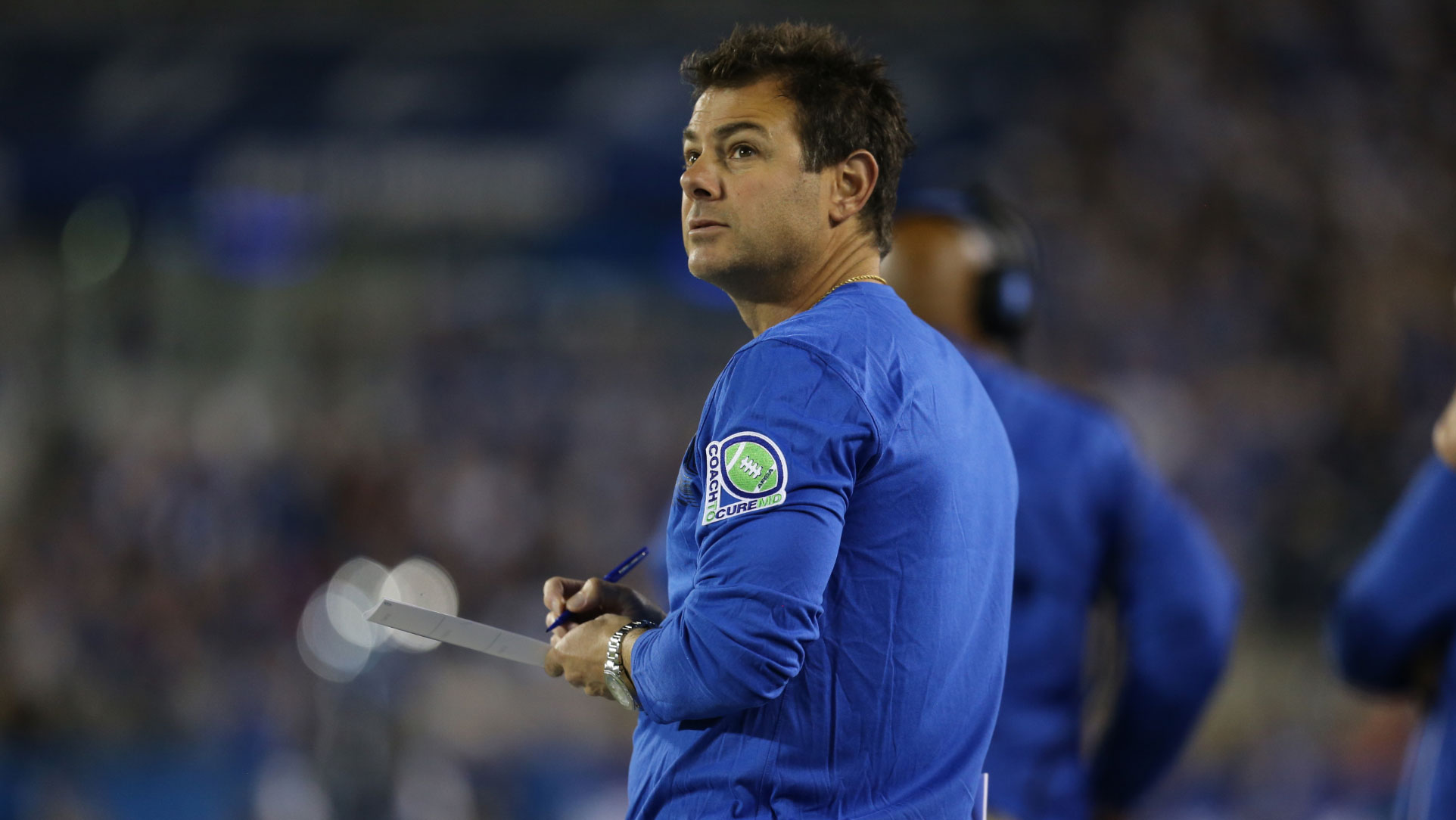 Frank Buffano, a middle-aged white man with brown hair, looks up and over his shoulder during a football game. He holds a clipboard and a pen and is wearing a blue UK coaching shirt.