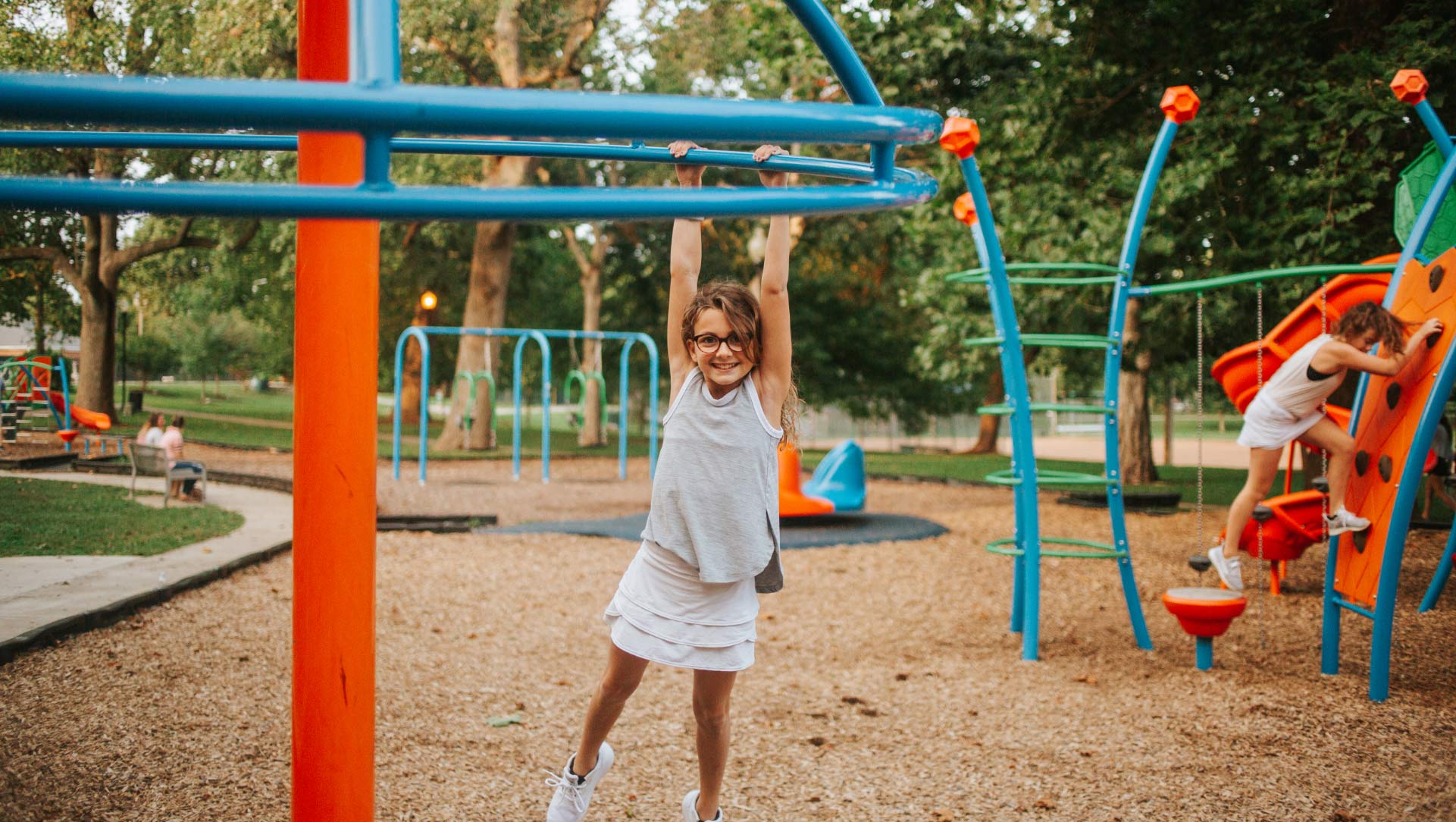 Gabriella Buffano, approximately 10 years old, holds onto a piece of playground equipment, swinging by her arms and grinning. She is wearing a white skirt, a gray tank top, and glasses.