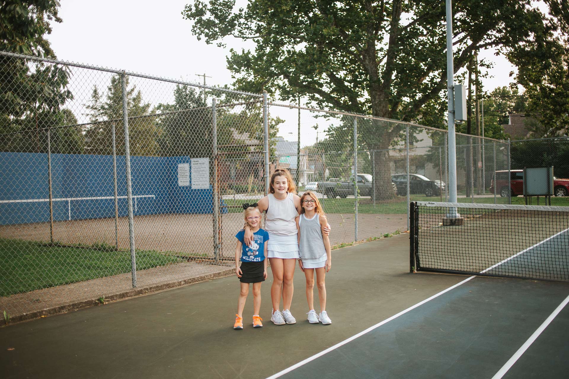 The Buffano sisters stand next to each other on a tennis court. Isabella, the oldest, has her arms around her younger sisters. All are smiling.