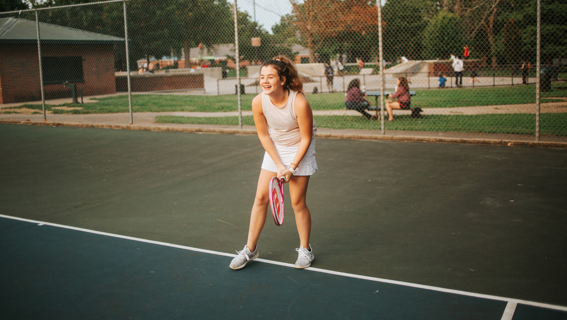 An action shot of Isabella, 12 years old, on the tennis court. She is wearing a white skirt and a gray tank top, and her curly brown hair is pulled back inn a ponytail.