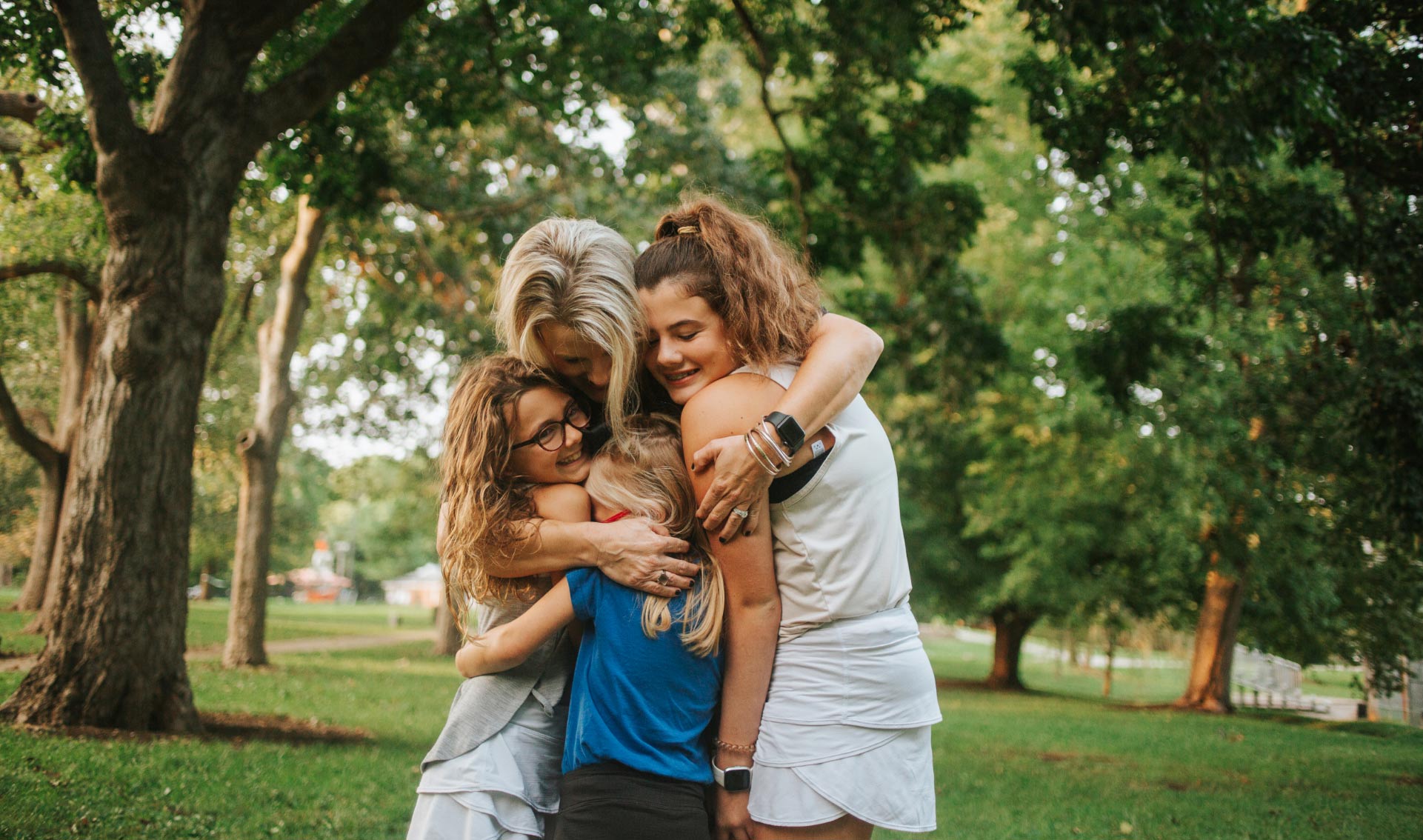 Kelley hugs her girls tightly. They are standing in a park. All are smiling.