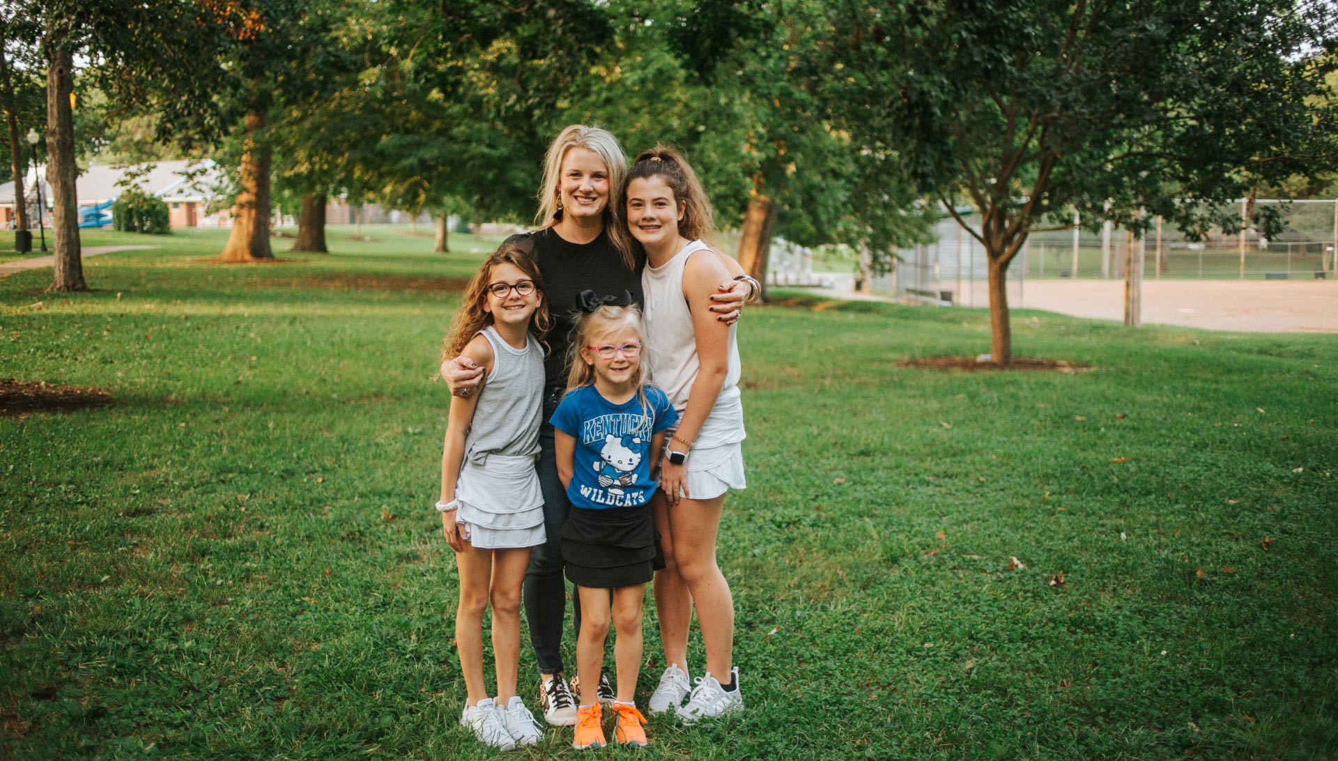 Kelley Buffano, a middle-aged white woman with blonde hair, stands in a park hugging her three daughters: Gabriellla, Olivia, and Isabella.