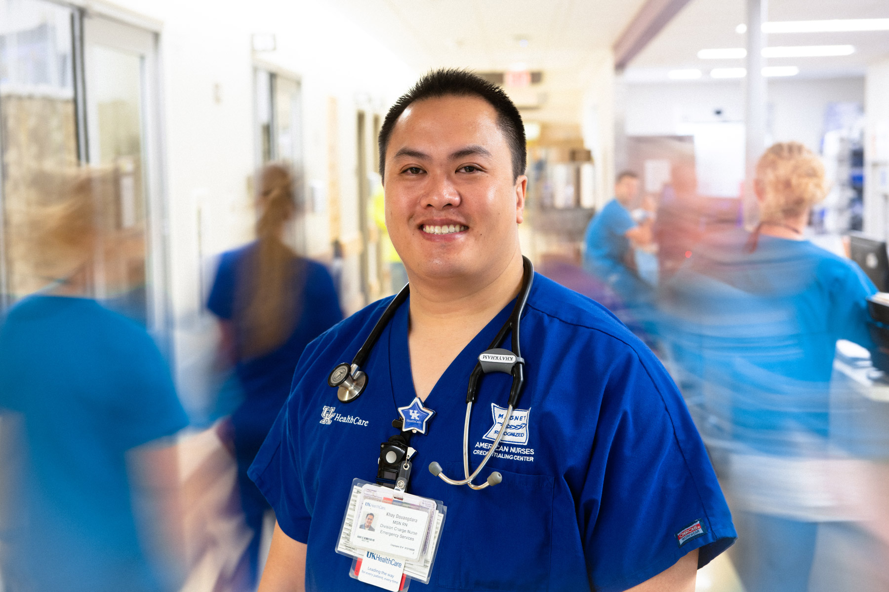 Khay smiles for a photo in his blue UK HealthCare nursing scrubs while other nurses are blurred walking past him.