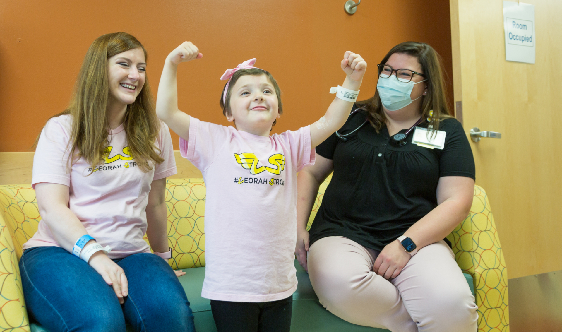 Leorah has a smile on her face and her arms in the air as she strikes a pose, while her mother Nicole and one of her nurses look on as they sit on a couch, smiling and laughing. Nicole is a white adult woman with light-brown hair and is wearing a pink “Leorah Strong” shirt. The nurse is a white adult woman with brown hair and is wearing a medical mask, a black half-button shirt, and khakis.