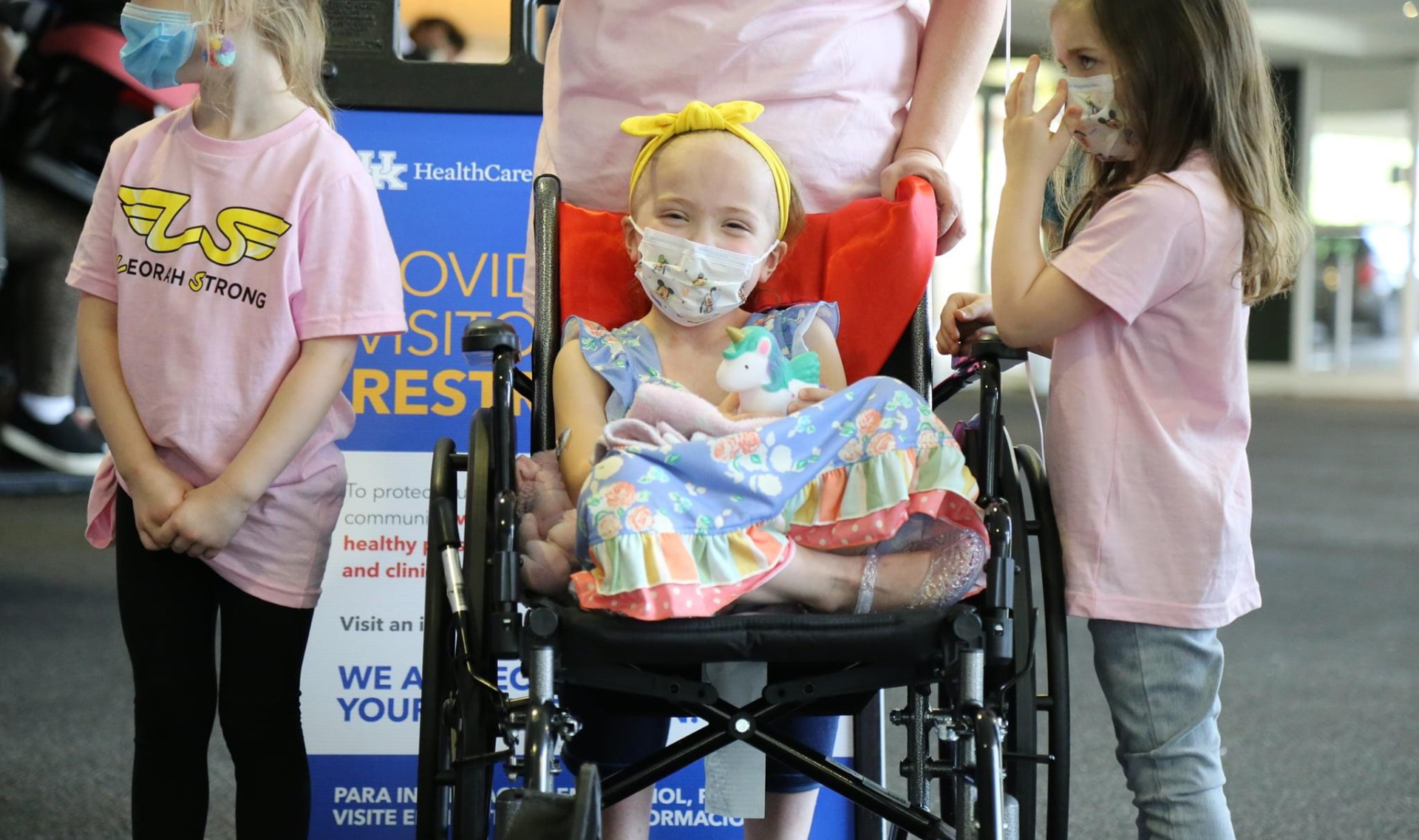 Leorah sits in her wheelchair, legs crossed and visibly smiling from behind her medical mask, with two young girls by her side. Both of these young girls are white, wearing pink “Lenorah Strong” t-shirts and jeans and both have medium-length hair, one blonde and one brunette.