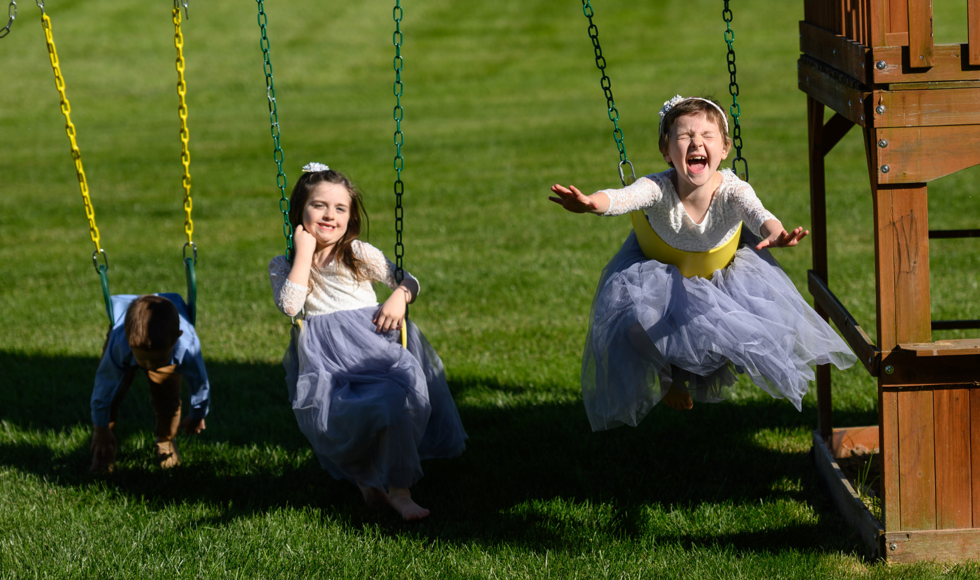 Leorah and her siblings play on their swingset, with Lenorah and her younger brother swinging while laying on their stomachs. Her brother is a young white boy, younger than his sisters, and is wearing a blue button-up and brown slacks. Her sister is a young white girl with brown hair, and is wearing a dress made of lace at the top and light blue tulle at the bottom.