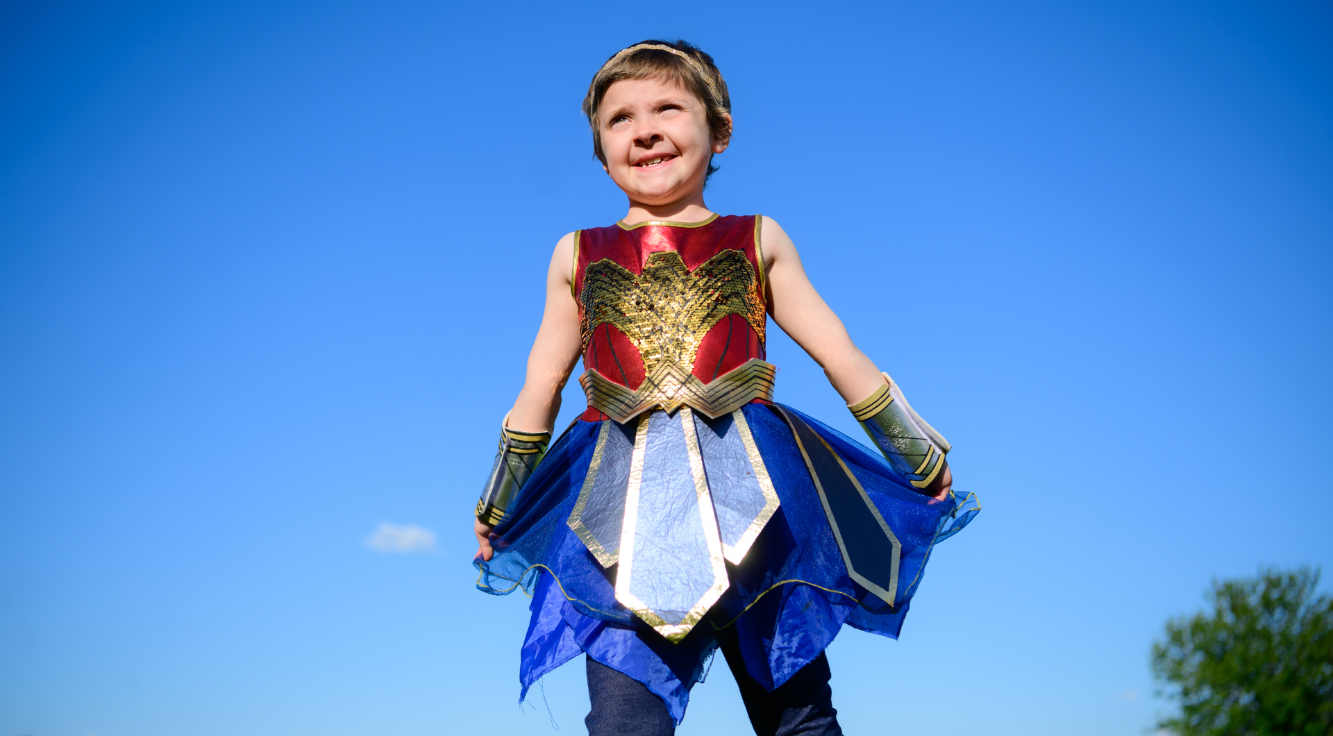 Leorah, a young white girl with short light brown hair, smiles as she stands in a field while wearing a Wonder Woman costume.