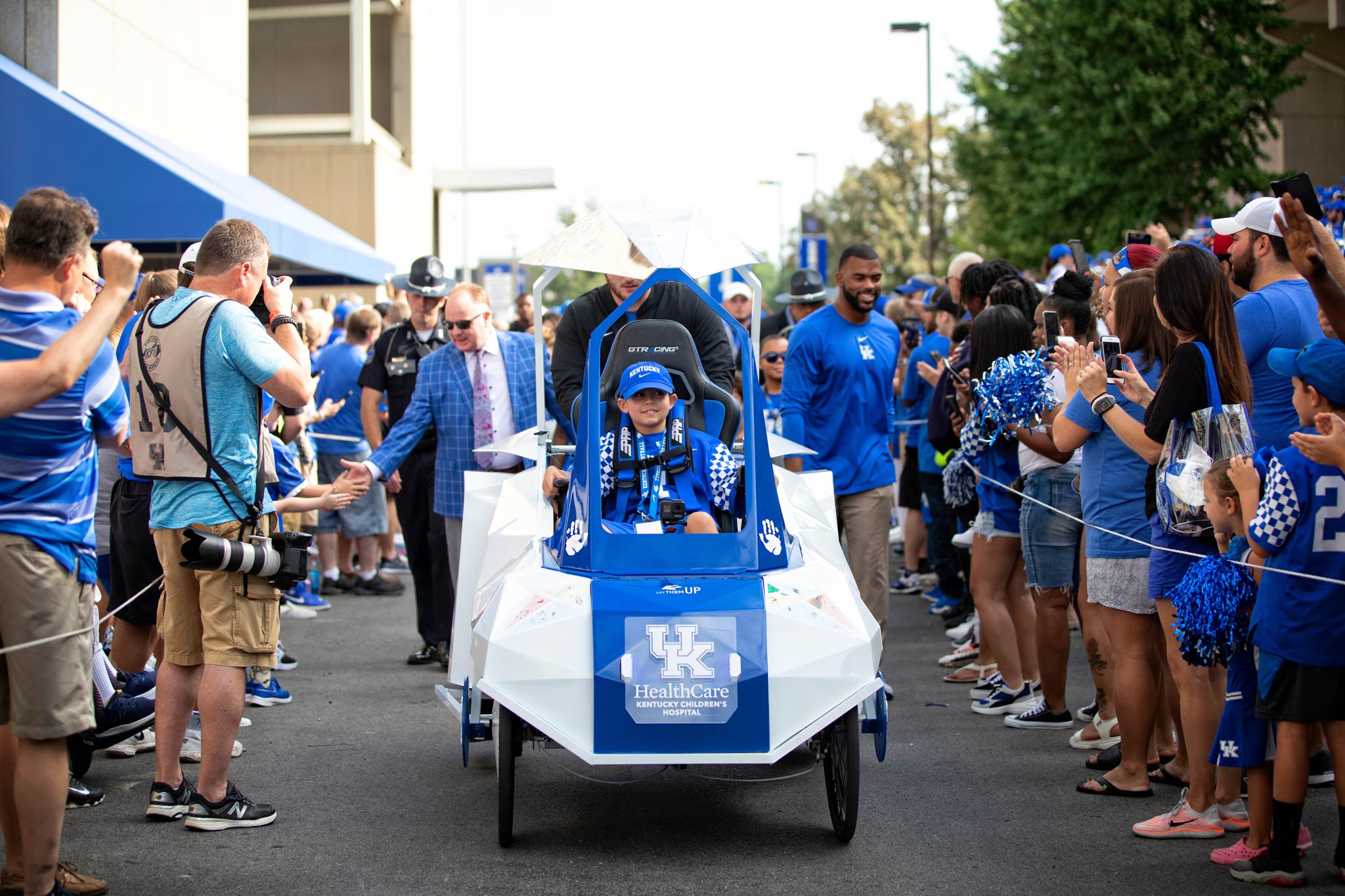 A little boy, riding in a futuristic-looking cart, leads Coach Stoops, Courtney Love, and the football team through a crowd of fans.